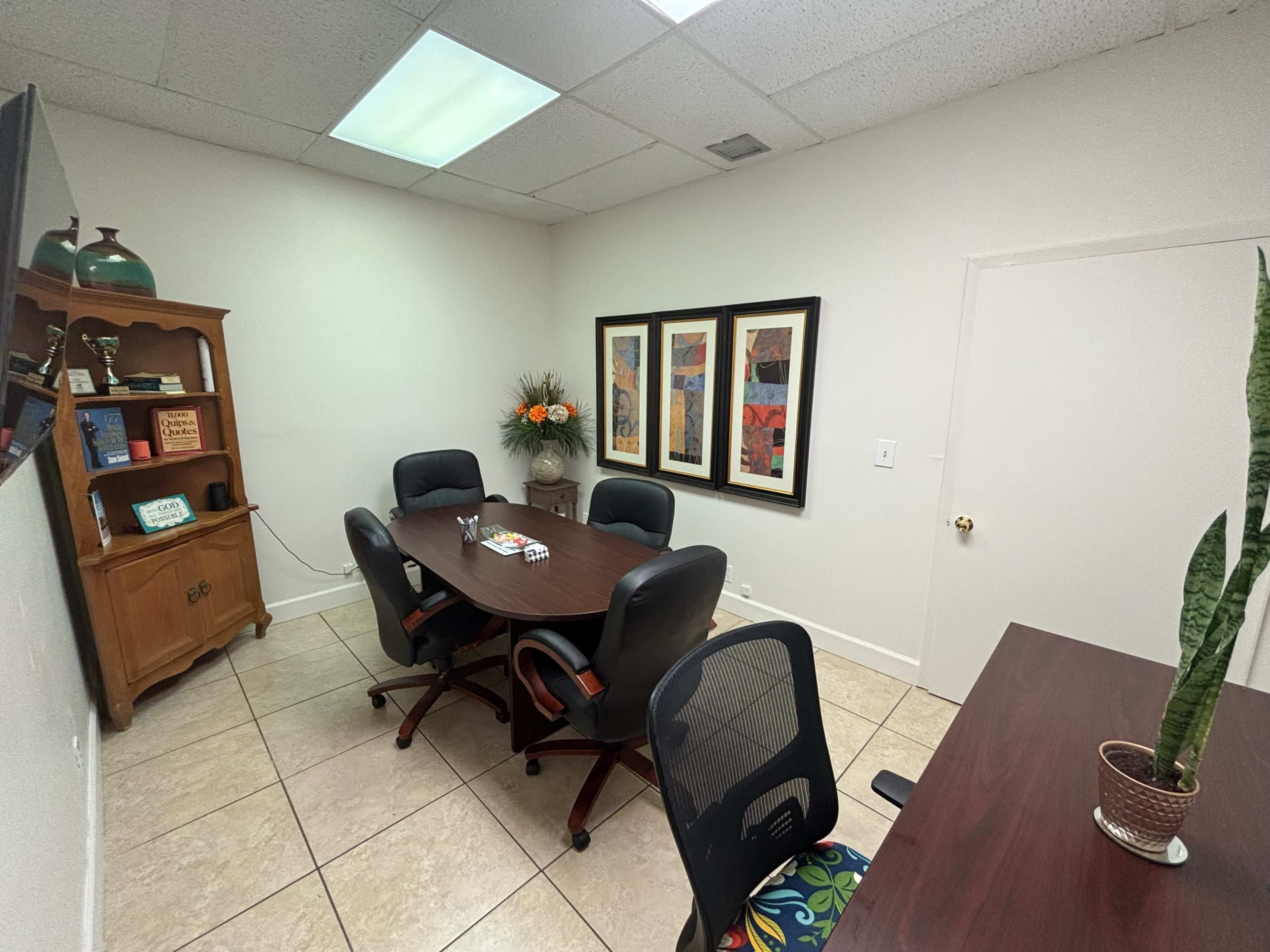 The image shows a small conference room with a round table surrounded by six chairs, a cabinet, and framed artwork on the wall.