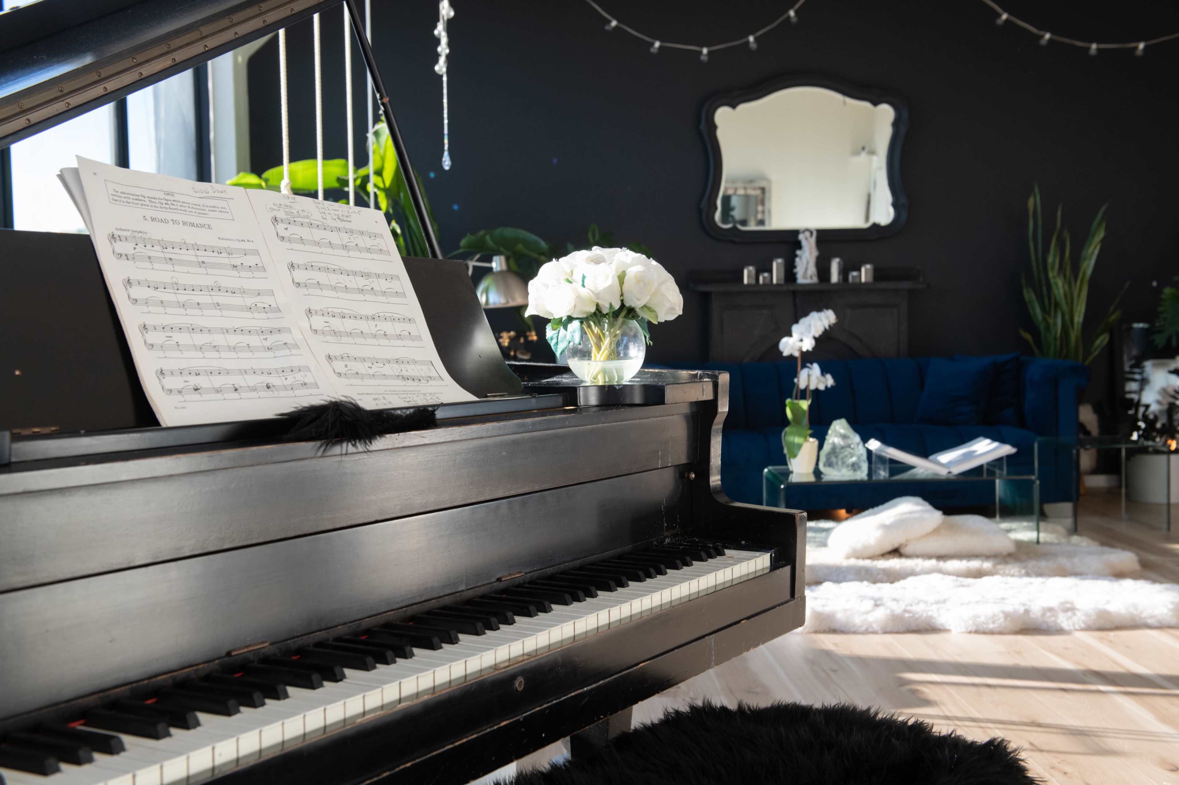 A black piano with sheet music sits in a bright room featuring a large mirror, a blue couch, and various plants.