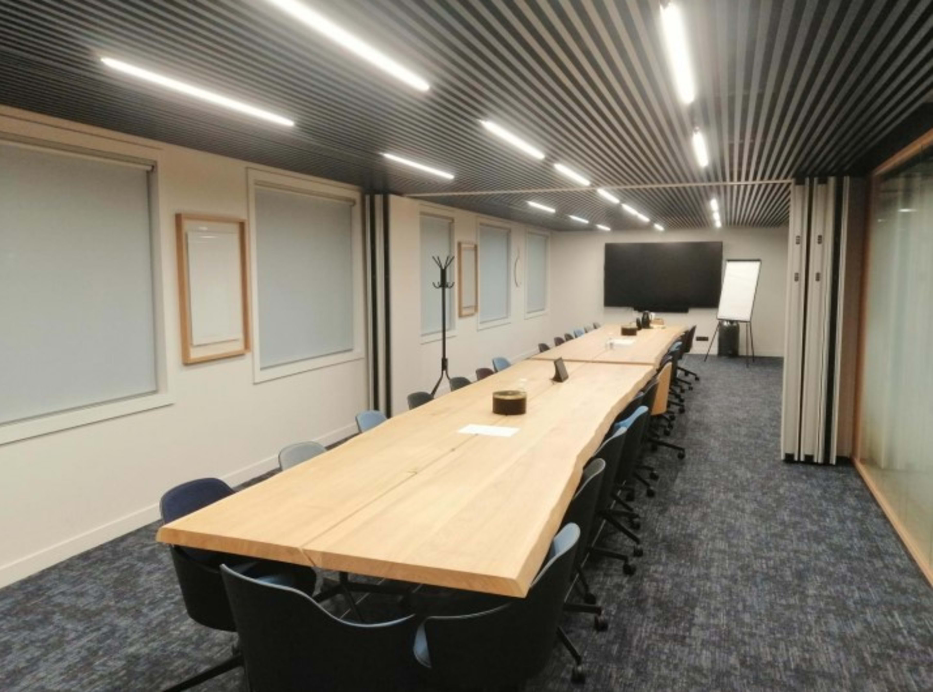 The image shows a modern conference room with a long wooden table, multiple chairs, and a large screen at one end.