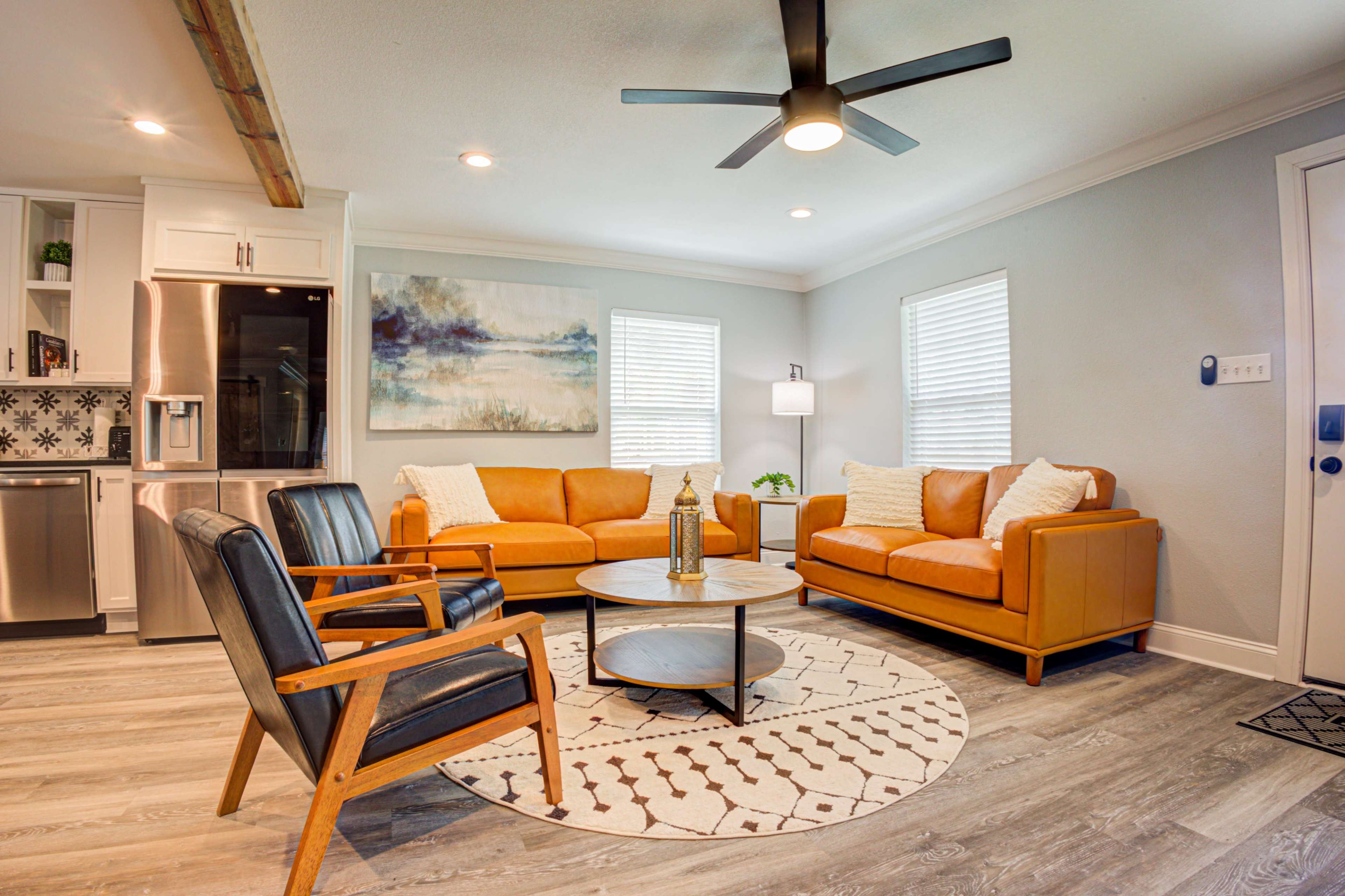 A modern living room with two orange sofas, a round coffee table, and black armchairs, featuring a ceiling fan and light-colored walls.