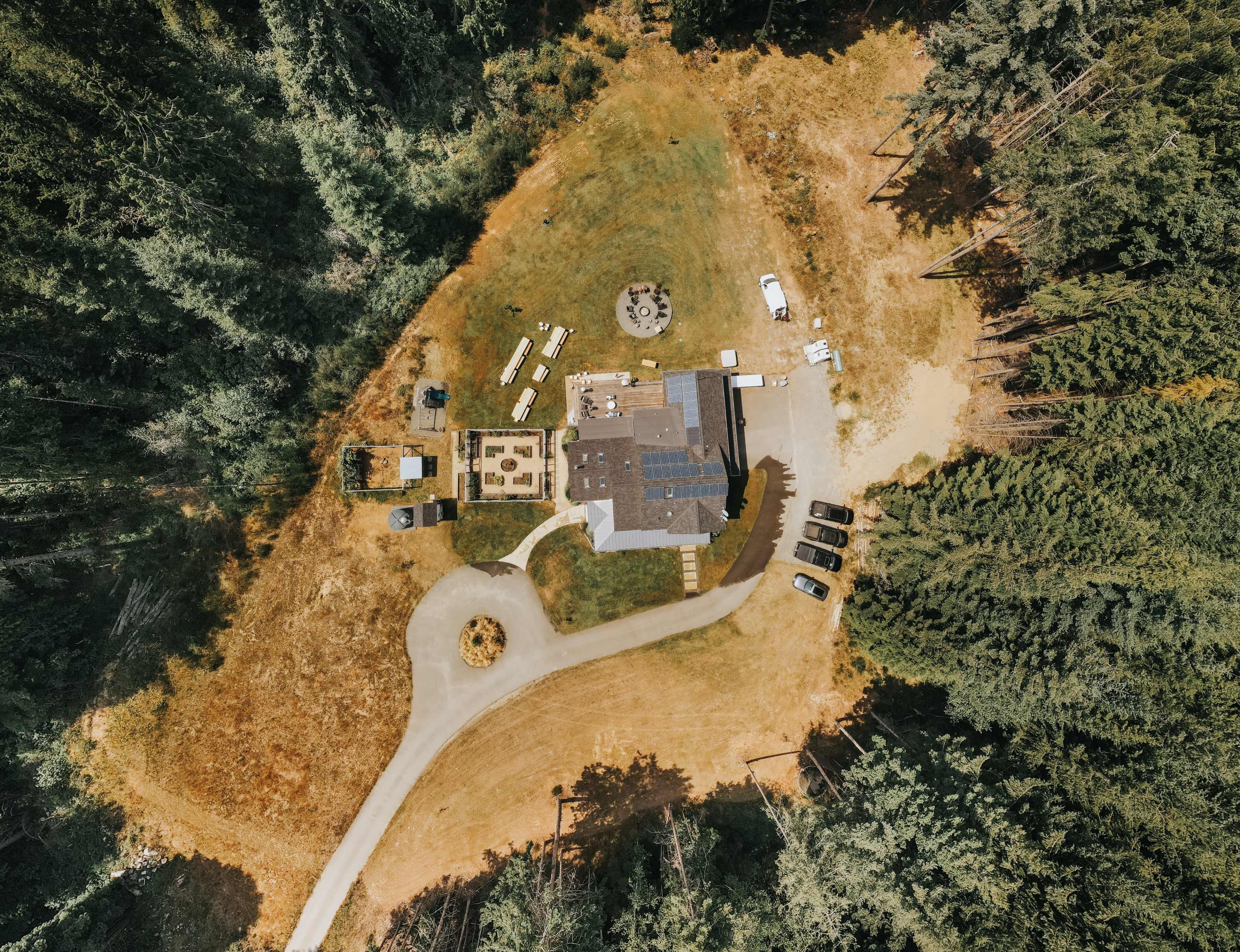 An aerial view of a house surrounded by trees, featuring a circular driveway, multiple parked vehicles, and an outdoor seating area.