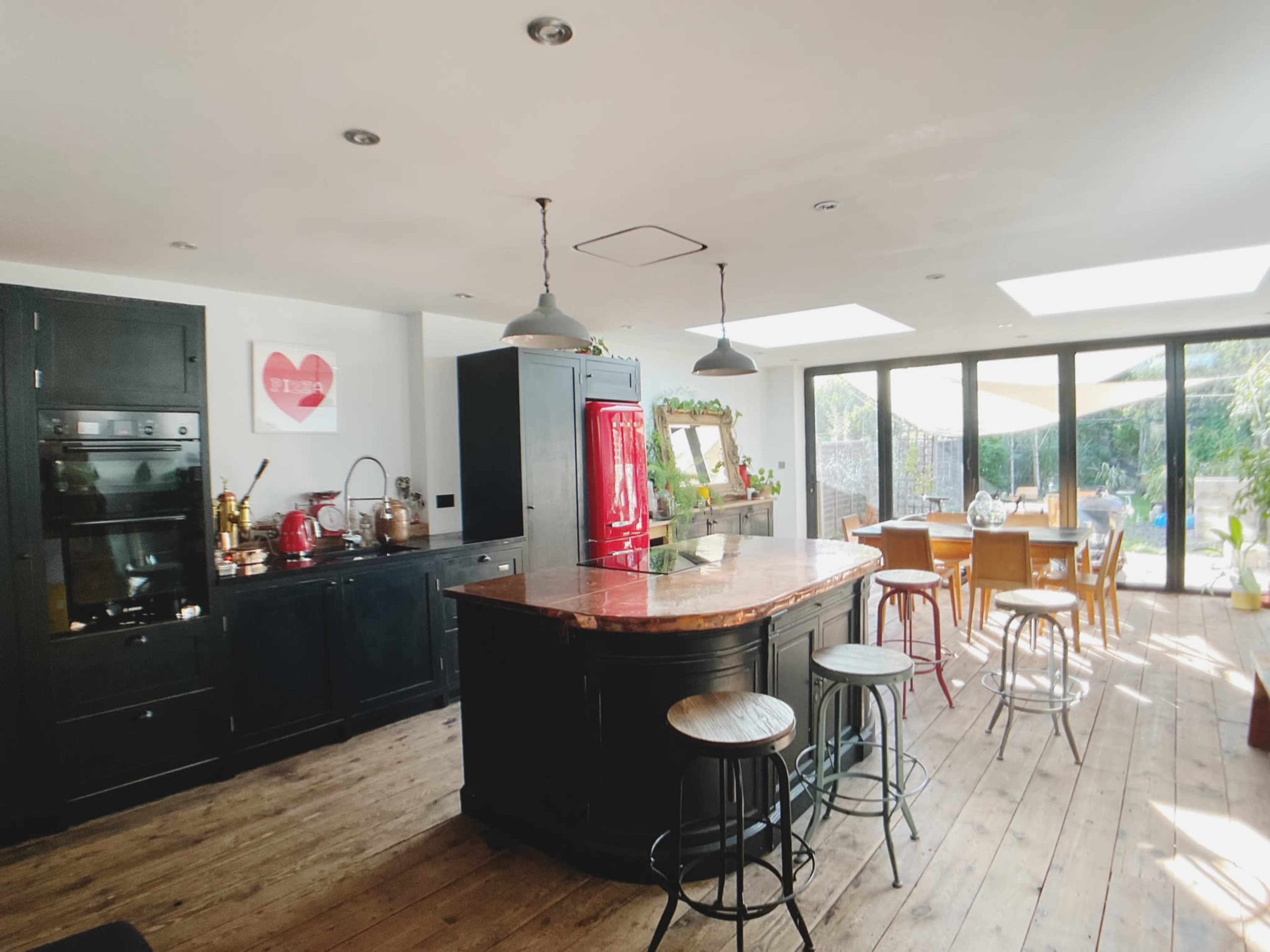 The kitchen features dark cabinetry, a large circular island with a copper countertop, and a dining area with wooden furniture illuminated by natural light from large windows.