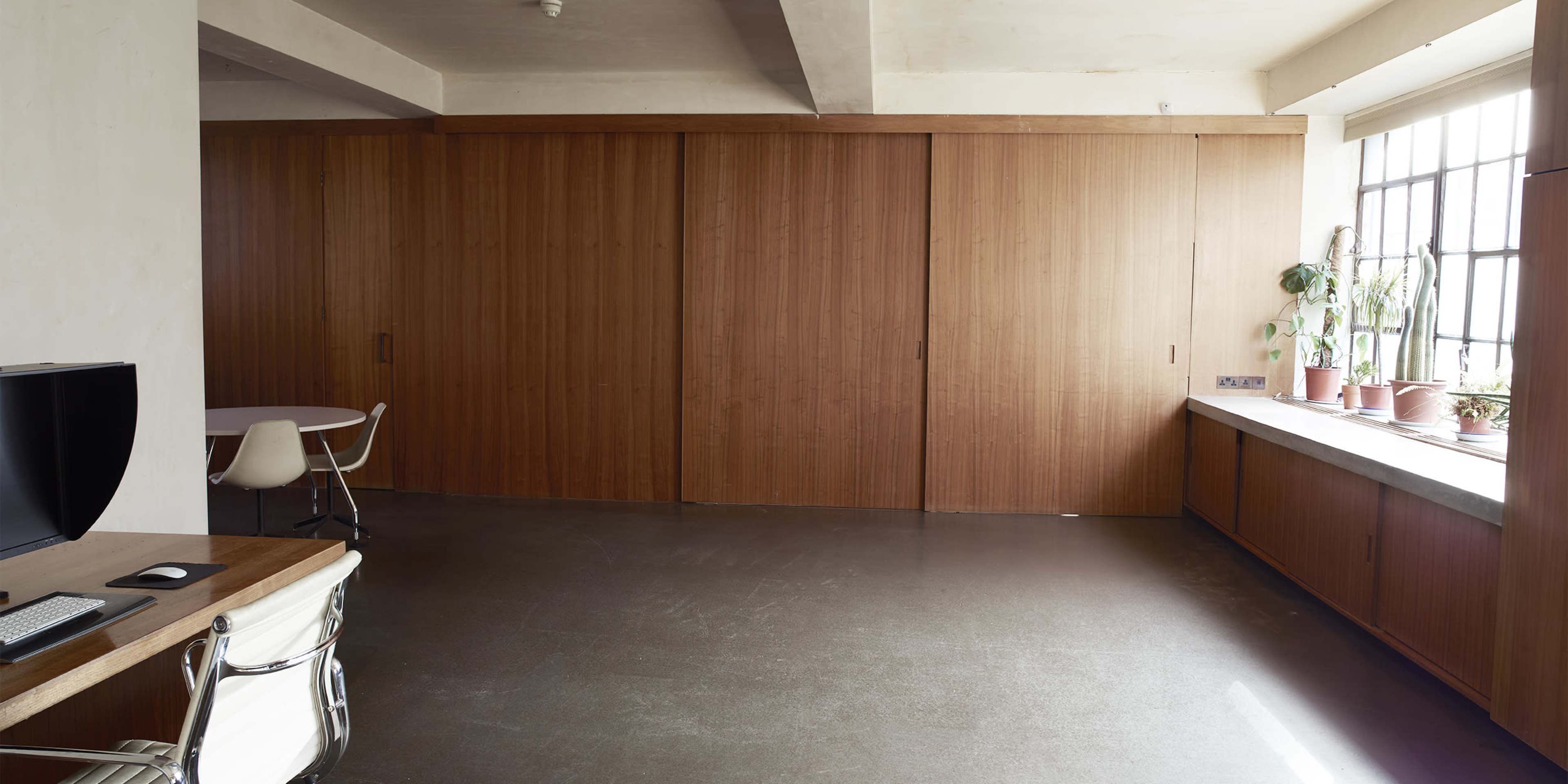 The image shows a minimalist interior space with wooden paneling, featuring a table and chairs on one side and large windows with potted plants on another side.
