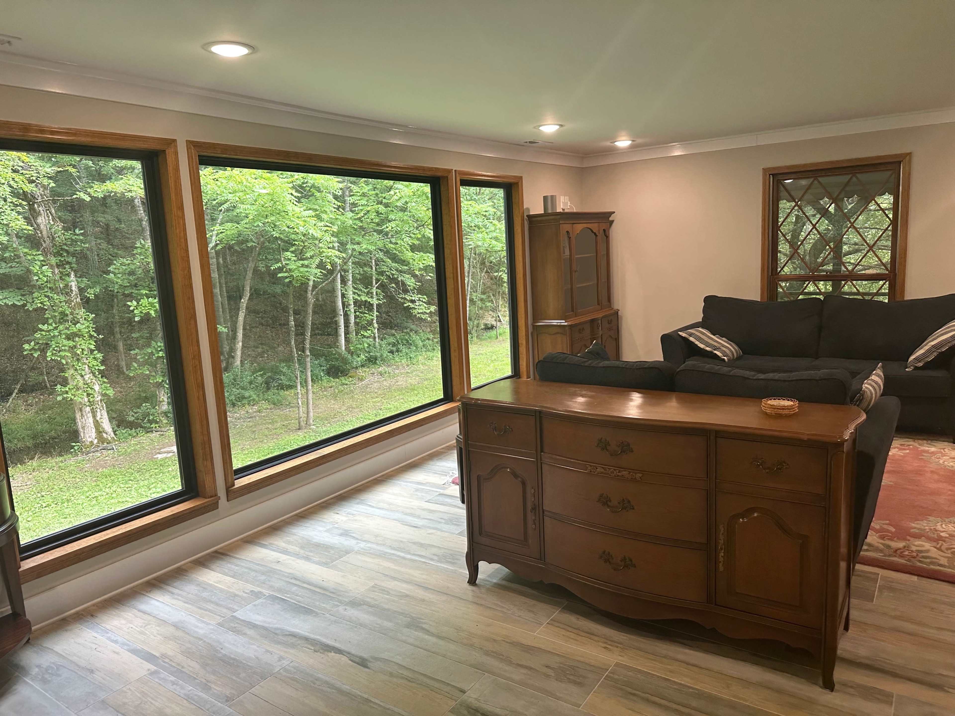 A wooden dresser sits in front of large windows that overlook a green, wooded area.