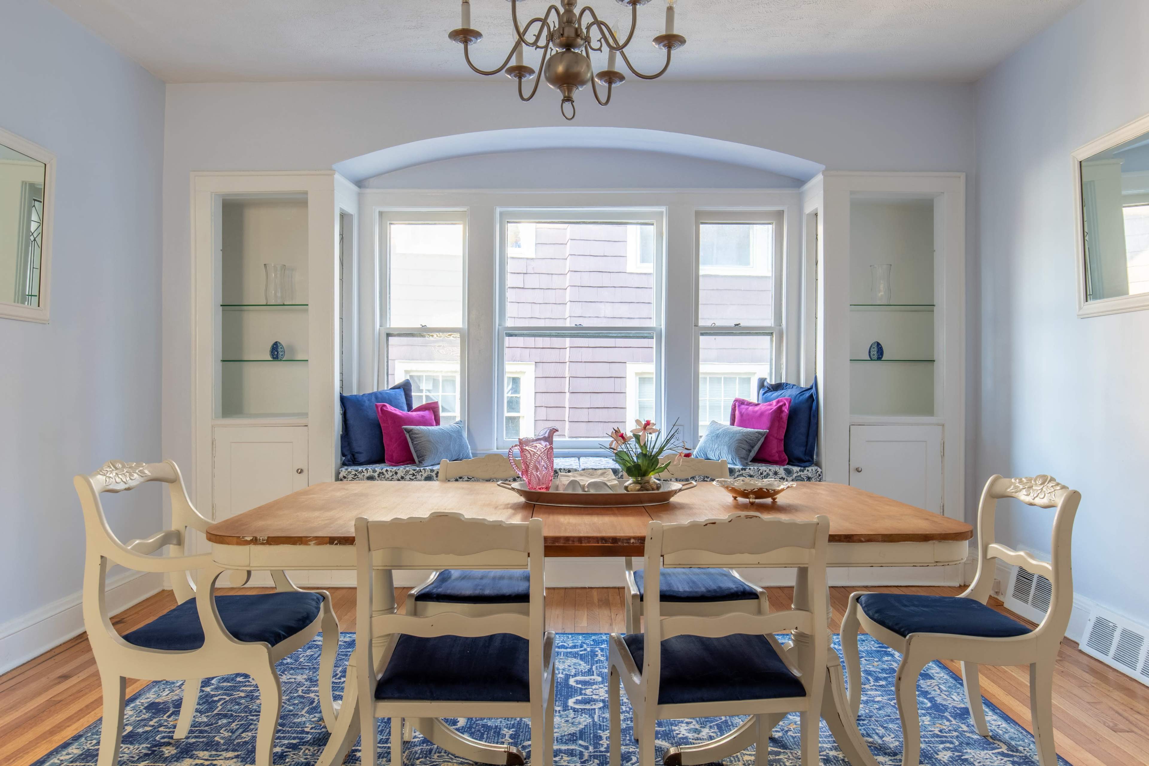 A dining room features a wooden table surrounded by six chairs, with a window nook on one side showcasing blue cushions and decorative elements.