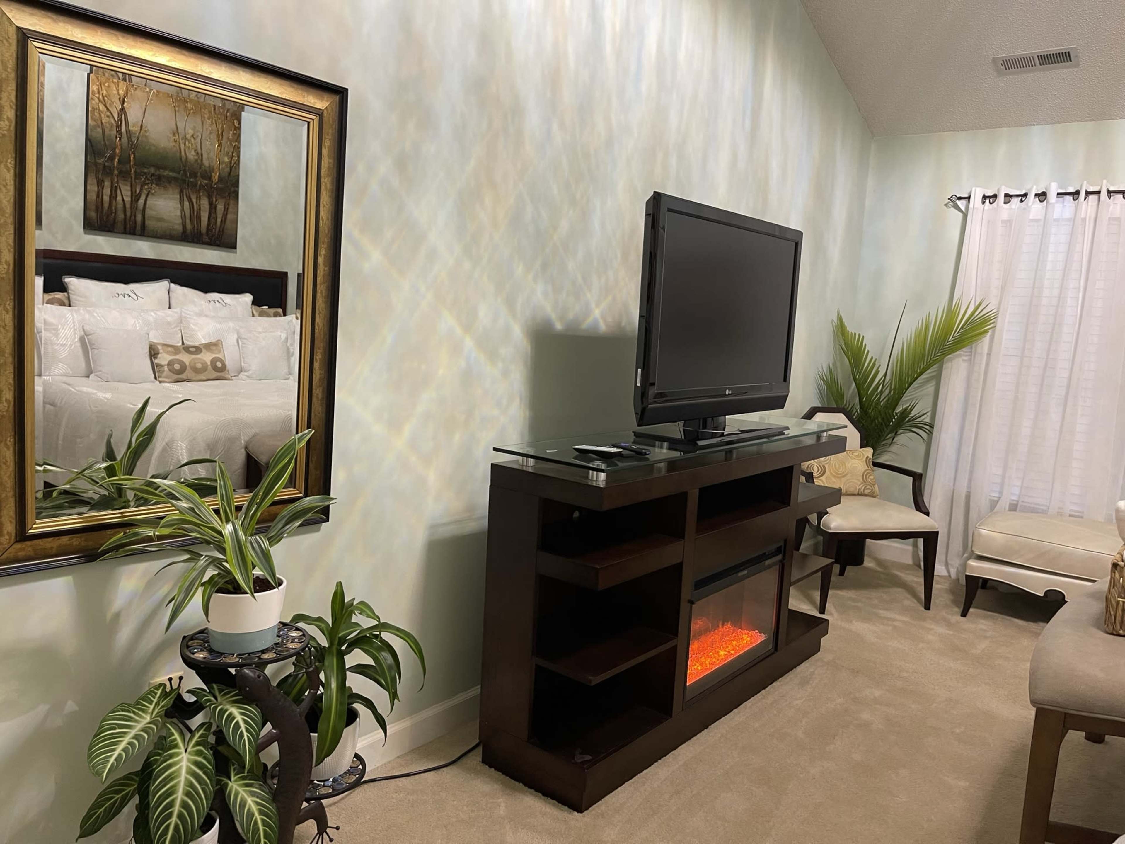 A cozy room featuring a television mounted on a dark wooden console with an electric fireplace, a mirror reflecting a bed in the background, and decorative plants beside a chair.