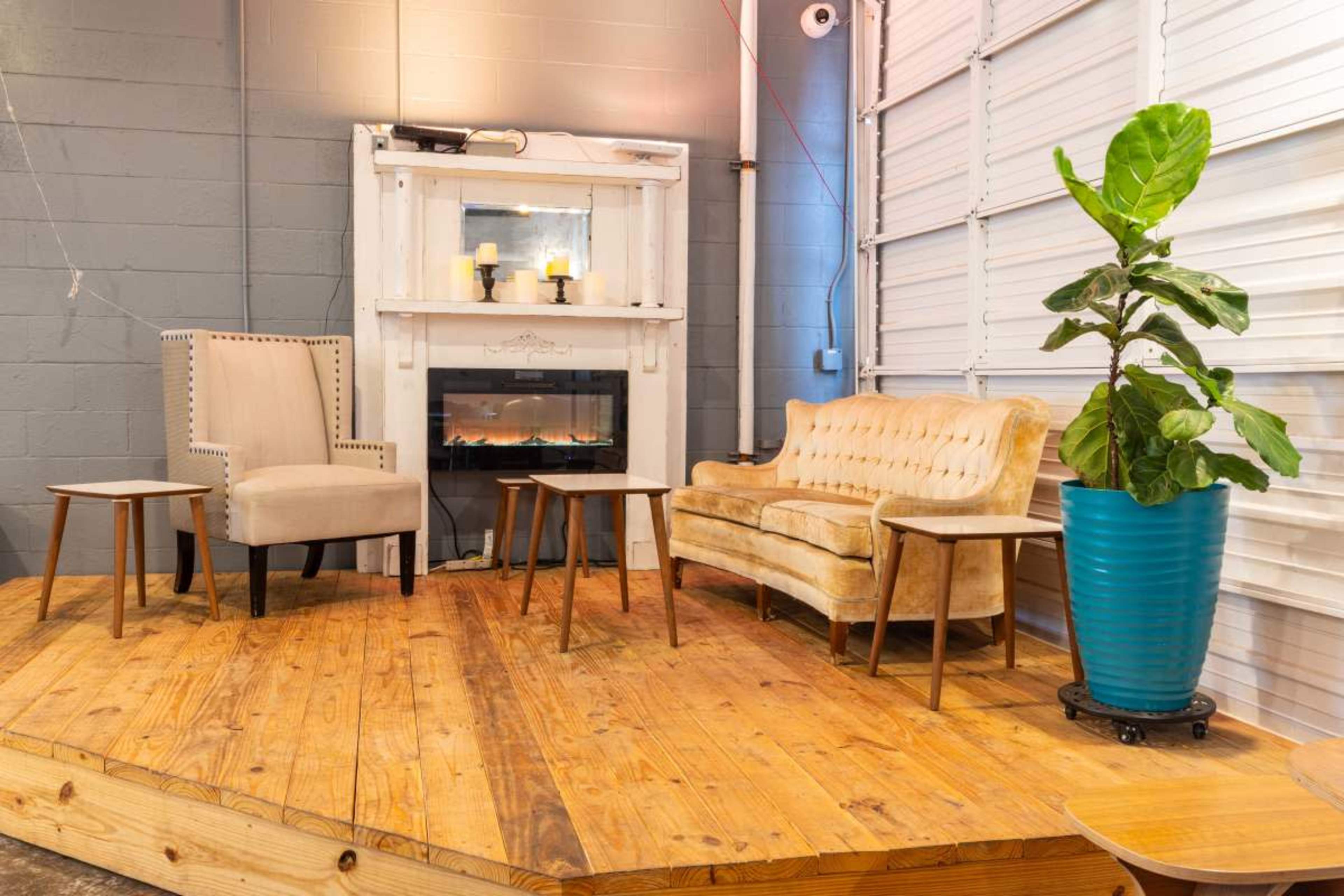 The image shows a cozy seating area with a beige sofa, a light-colored armchair, and three small tables, all set on a wooden platform next to a decorative fireplace and a large potted plant.