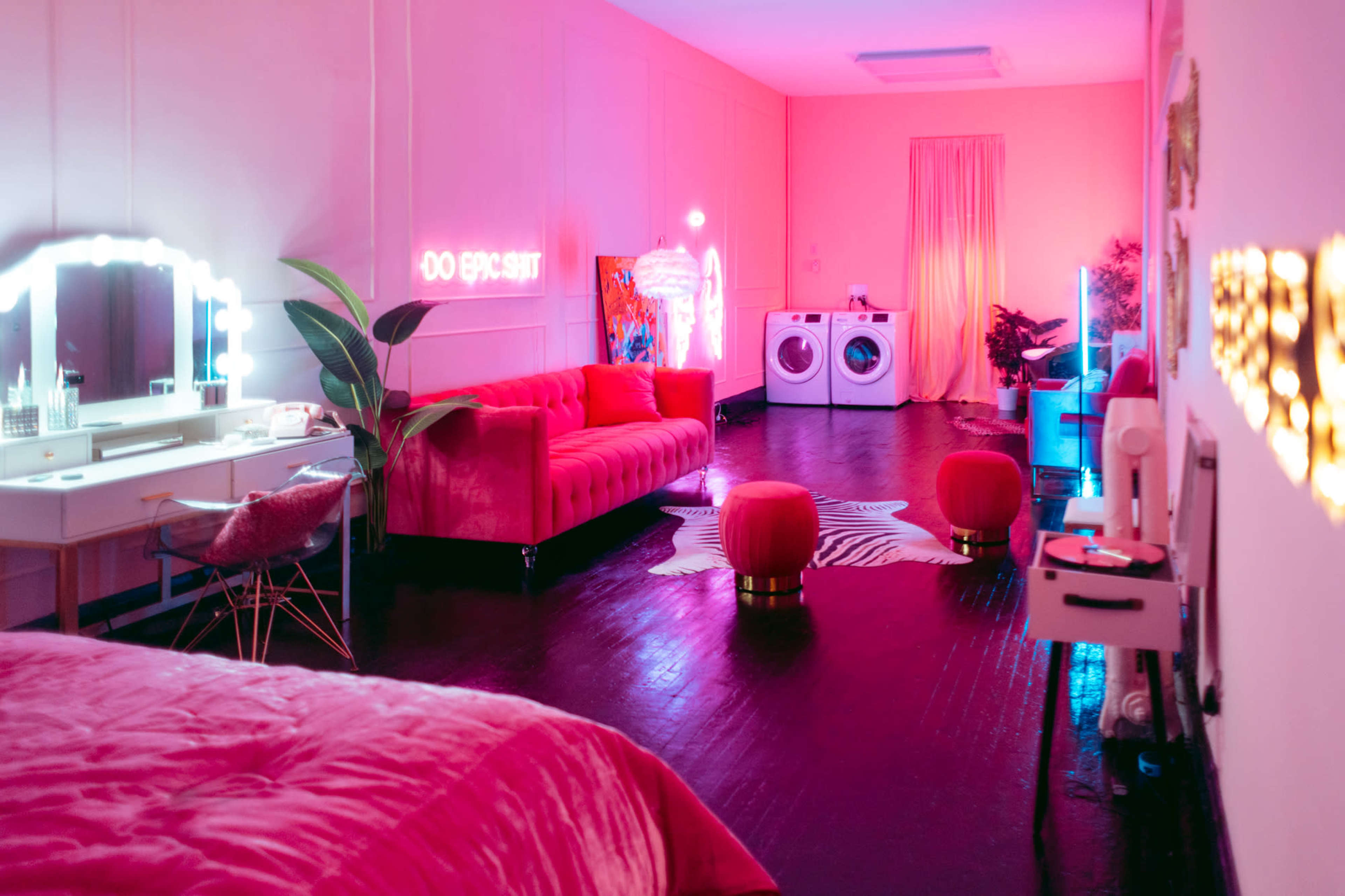 A brightly lit room features a pink couch, round poufs, a vanity area, and a washing machine, all set against a vibrant color scheme.