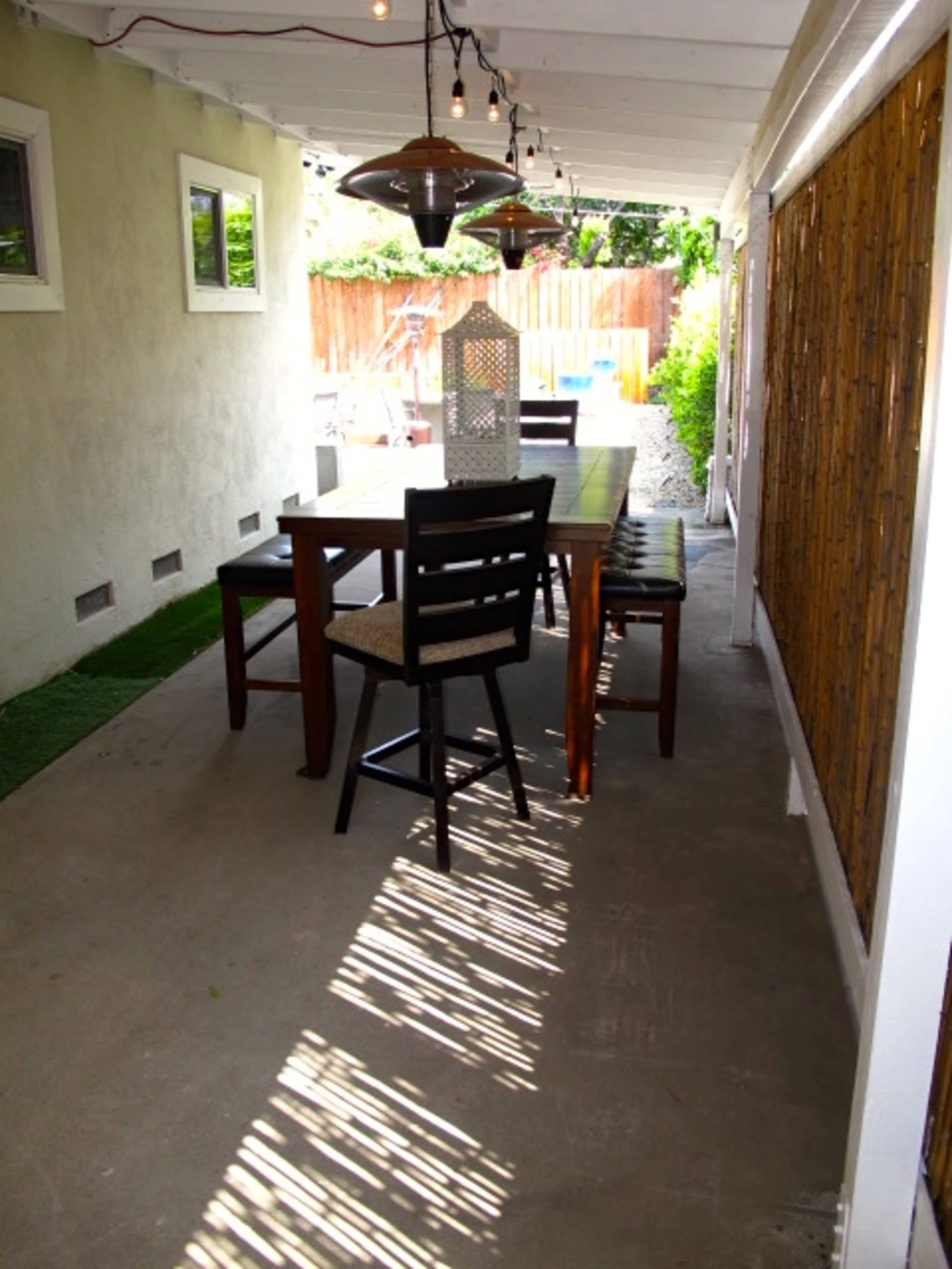 A patio area features a wooden dining table surrounded by chairs, with overhead lighting and greenery visible in the background.