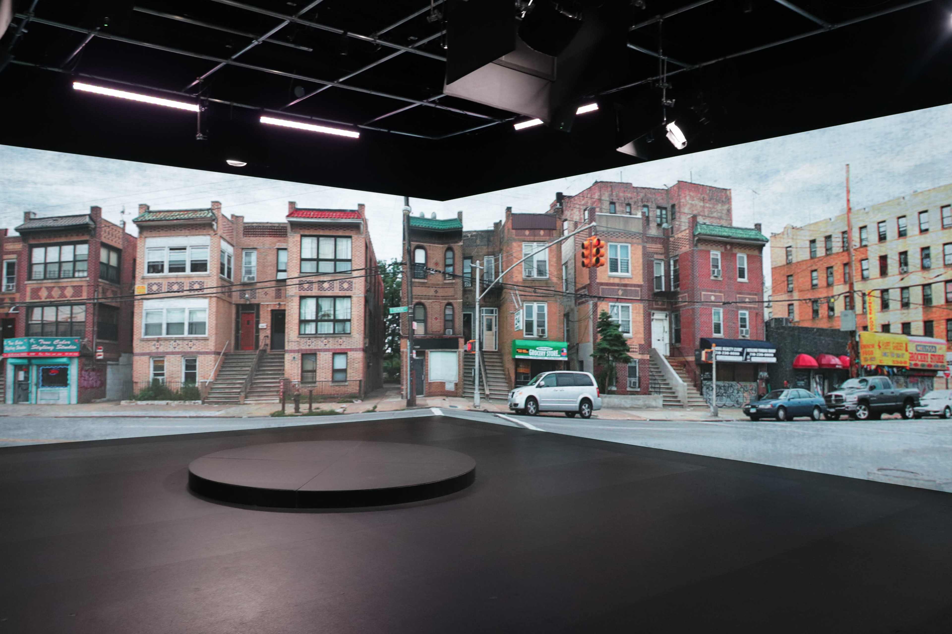 A studio with a circular stage features a backdrop of urban buildings and storefronts projected on the walls.