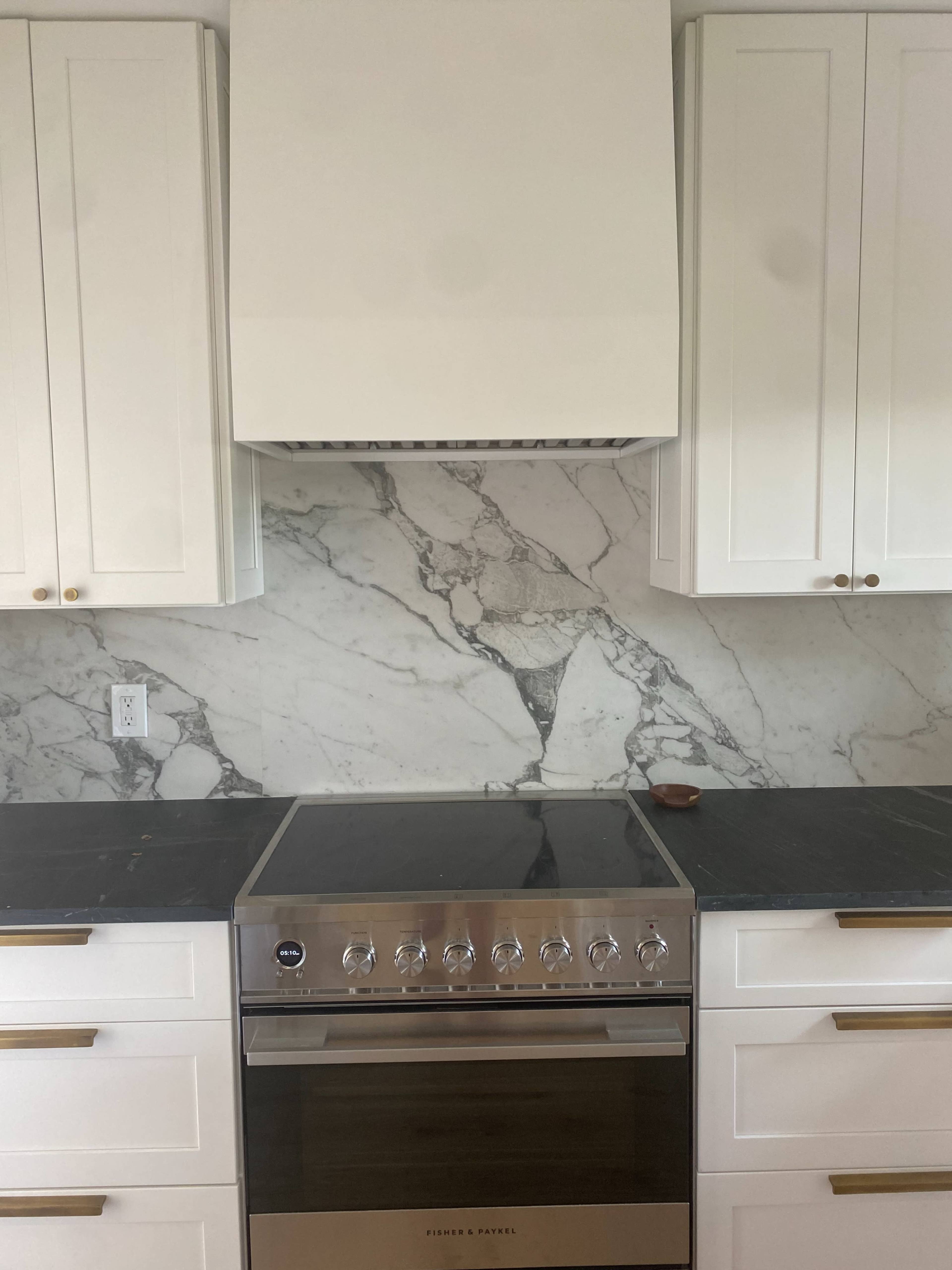 A stainless steel oven is installed beneath a white range hood, complemented by marble backsplash and dark countertops in a kitchen with white cabinetry.