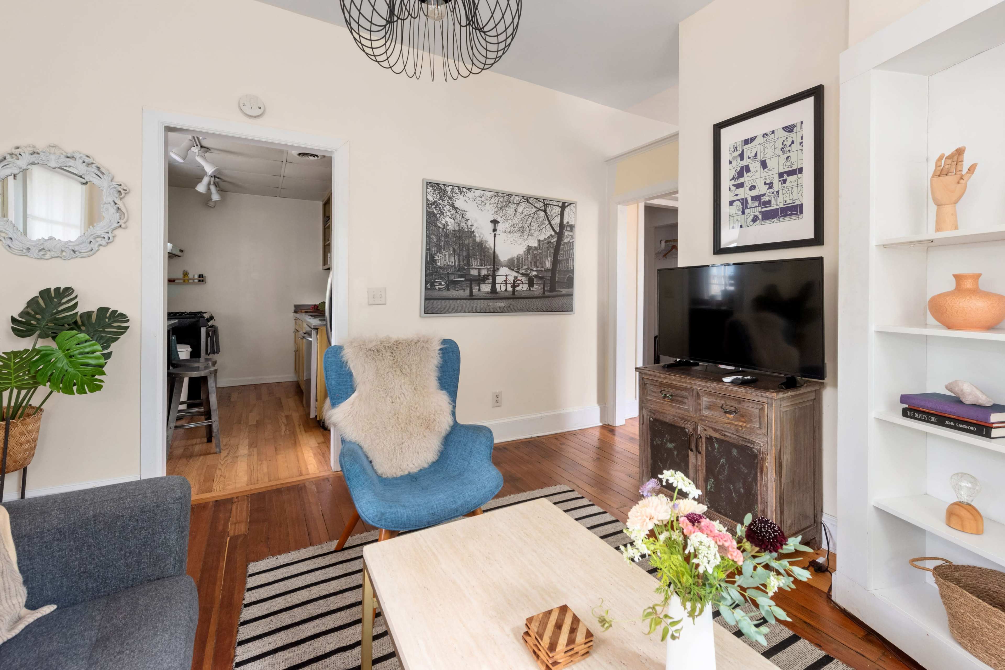 A cozy living room features a blue chair, a wooden coffee table with flowers, and a television on a rustic cabinet, with a glimpse of a kitchen in the background.