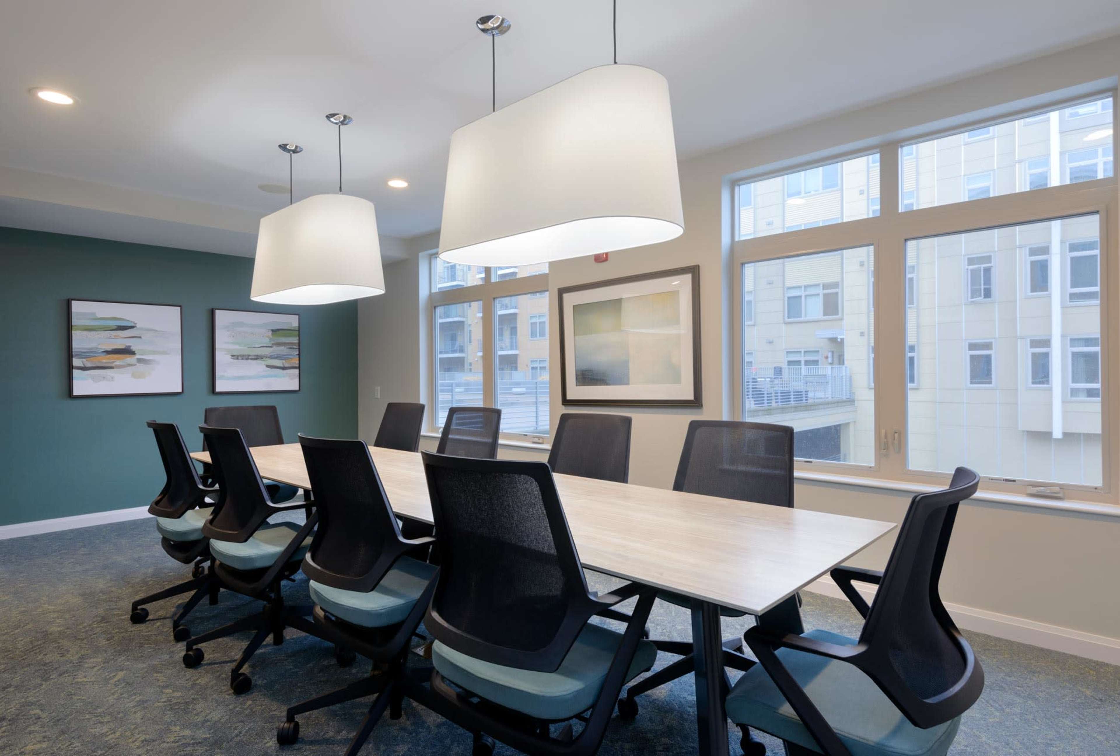 A modern conference room features a long table surrounded by eight ergonomic chairs, with large windows and two pendant lights overhead.