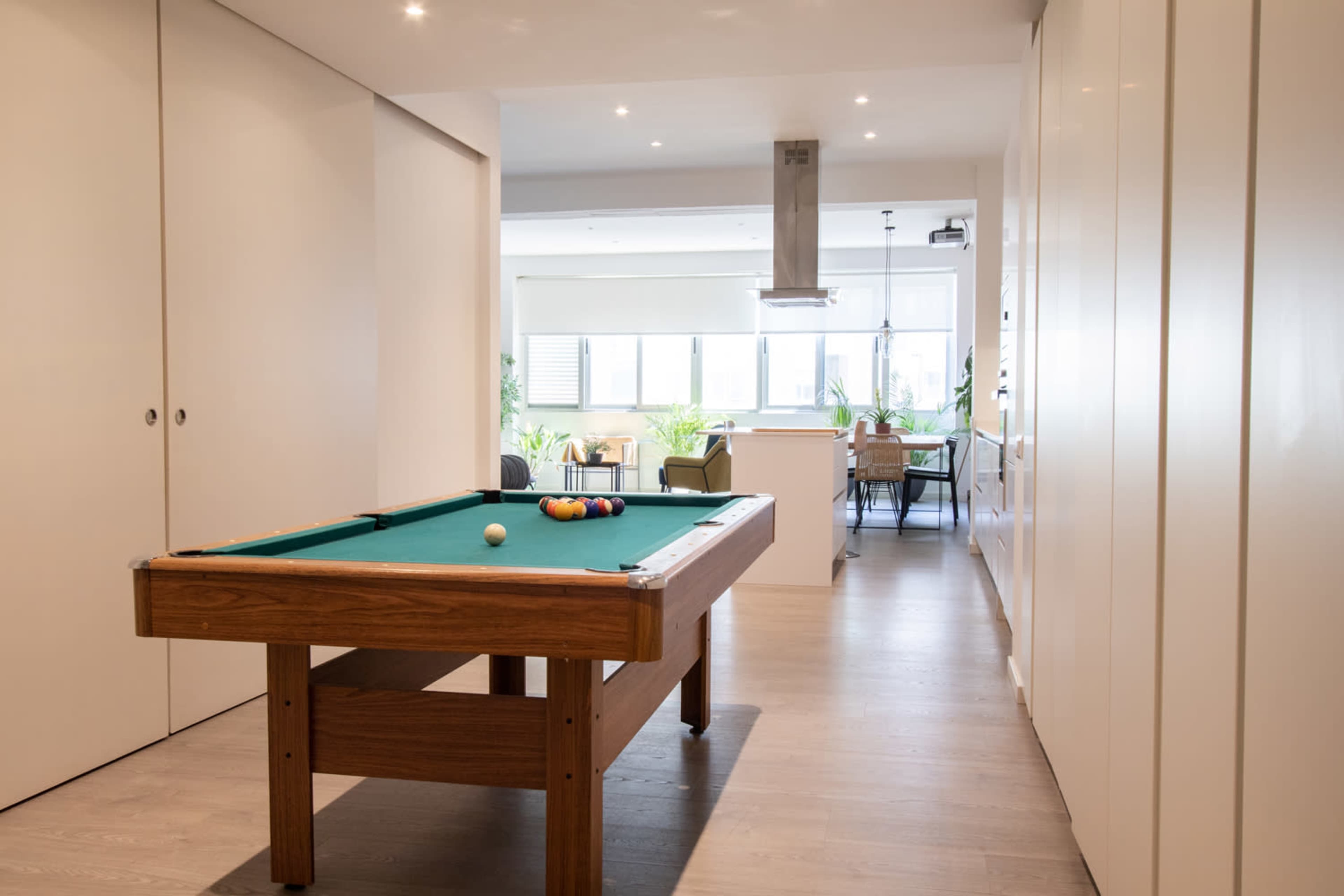 The image shows a modern interior space featuring a billiard table in the foreground and a dining area with plants in the background.