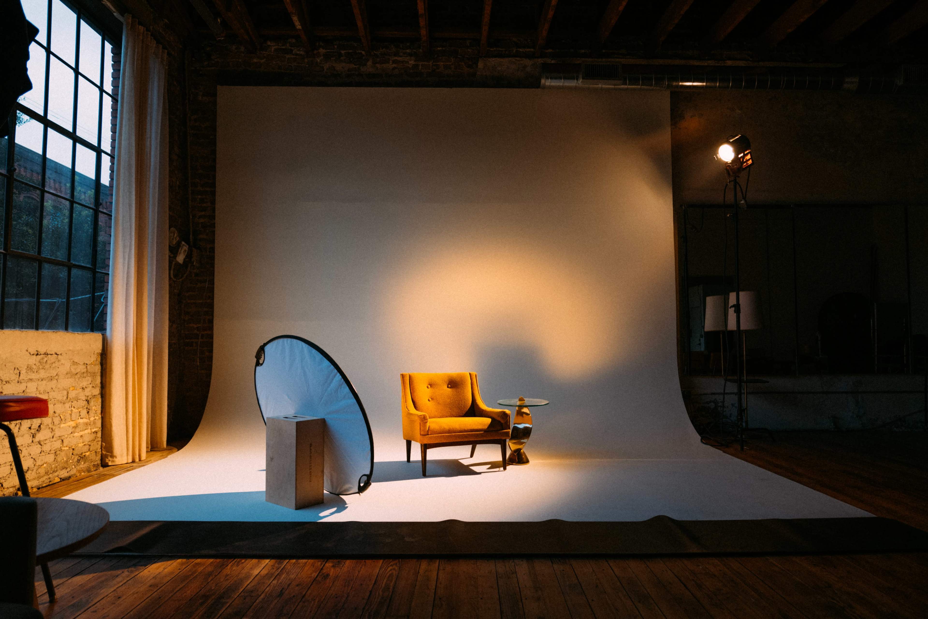 The image shows a yellow armchair positioned on a white backdrop in a studio, with a softbox light illuminating the scene and wooden flooring visible.
