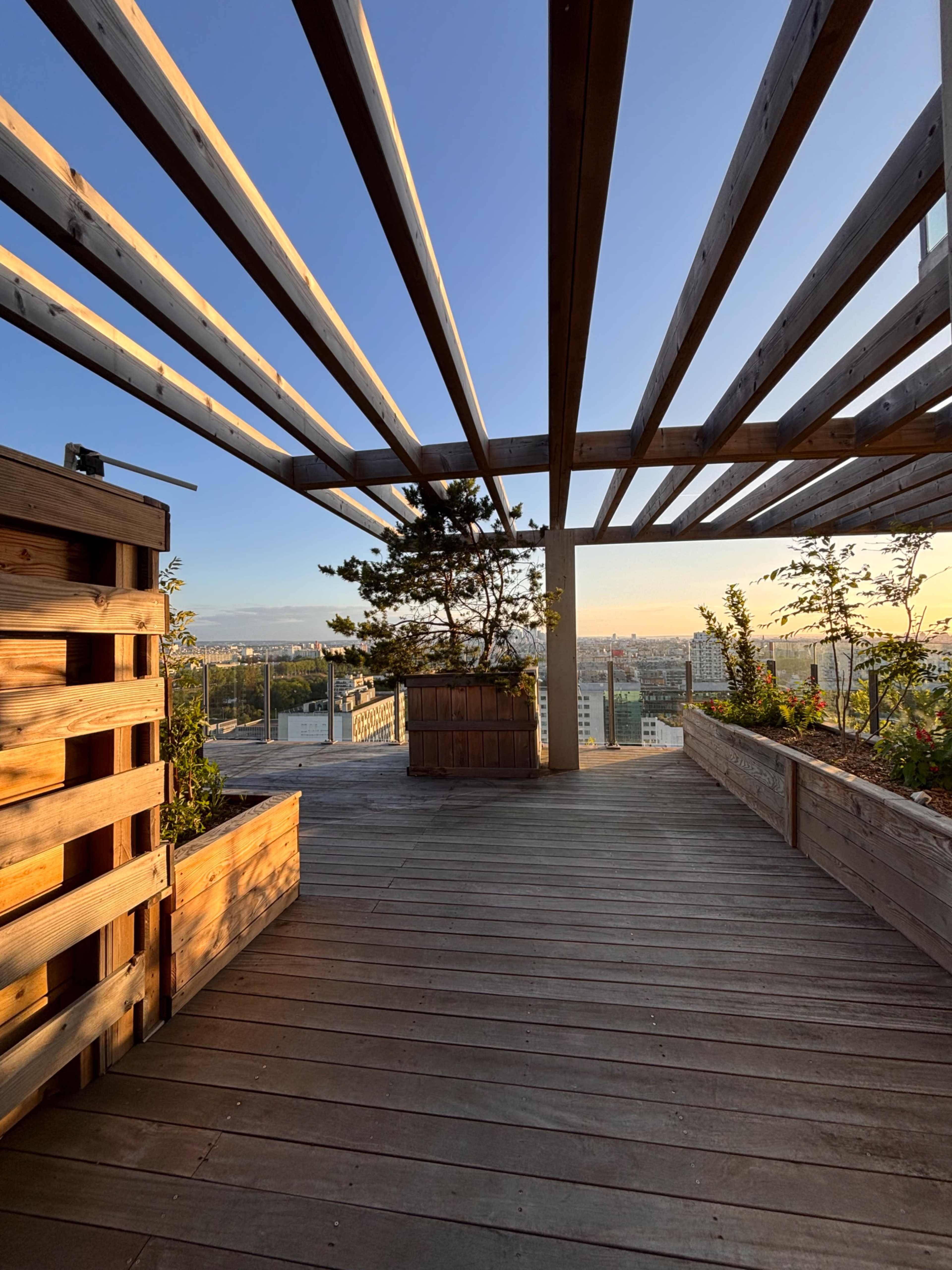 The image shows a wooden deck with a pergola overhead, overlooking a city skyline at sunset.