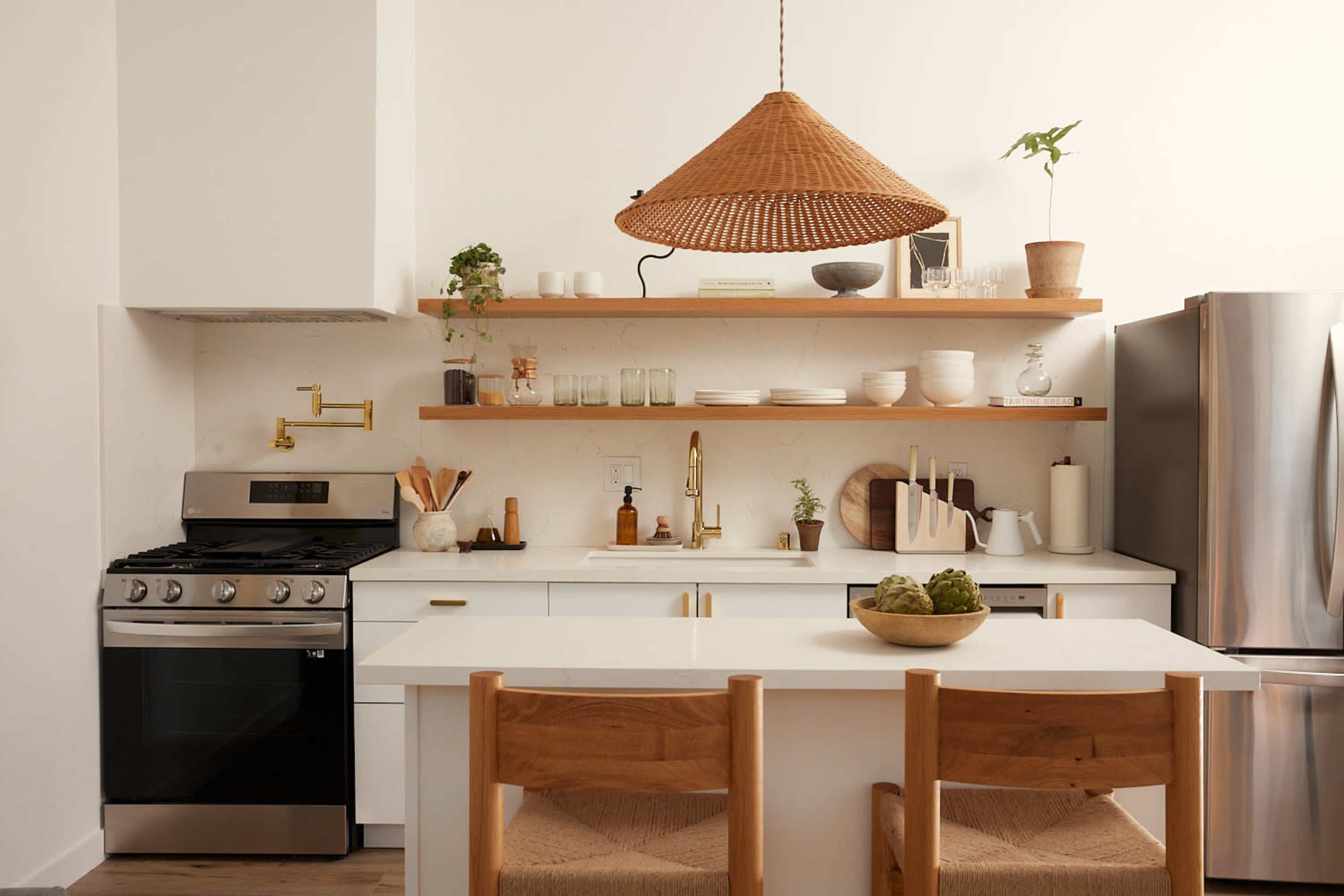 A modern kitchen features a gas stove, stainless steel refrigerator, wooden shelves with dishware, and a large pendant light hanging above a dining table with two wooden chairs.
