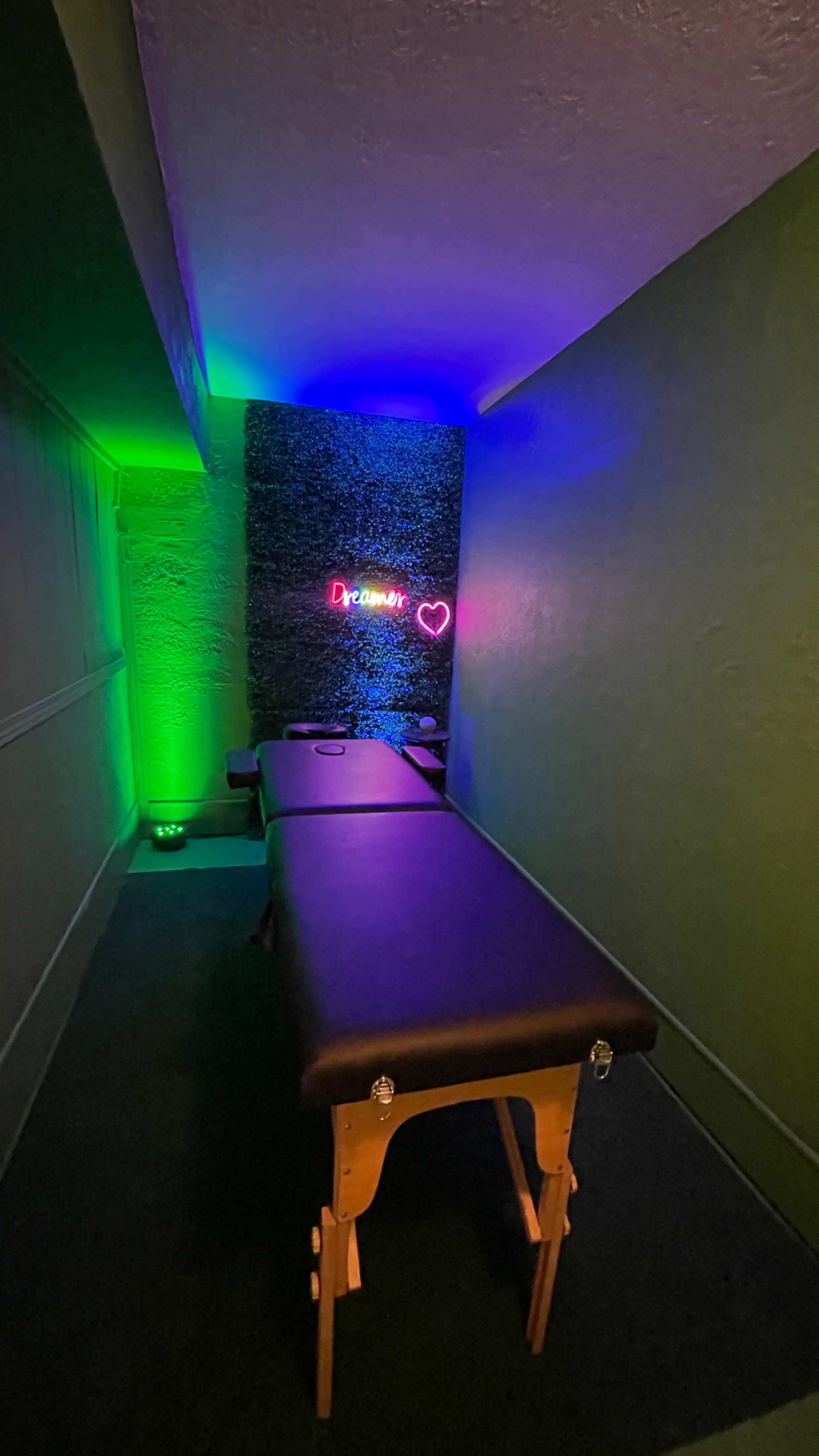 A massage room features a dark massage table under purple and green lighting, with a neon sign on the wall that reads "Dreaming."