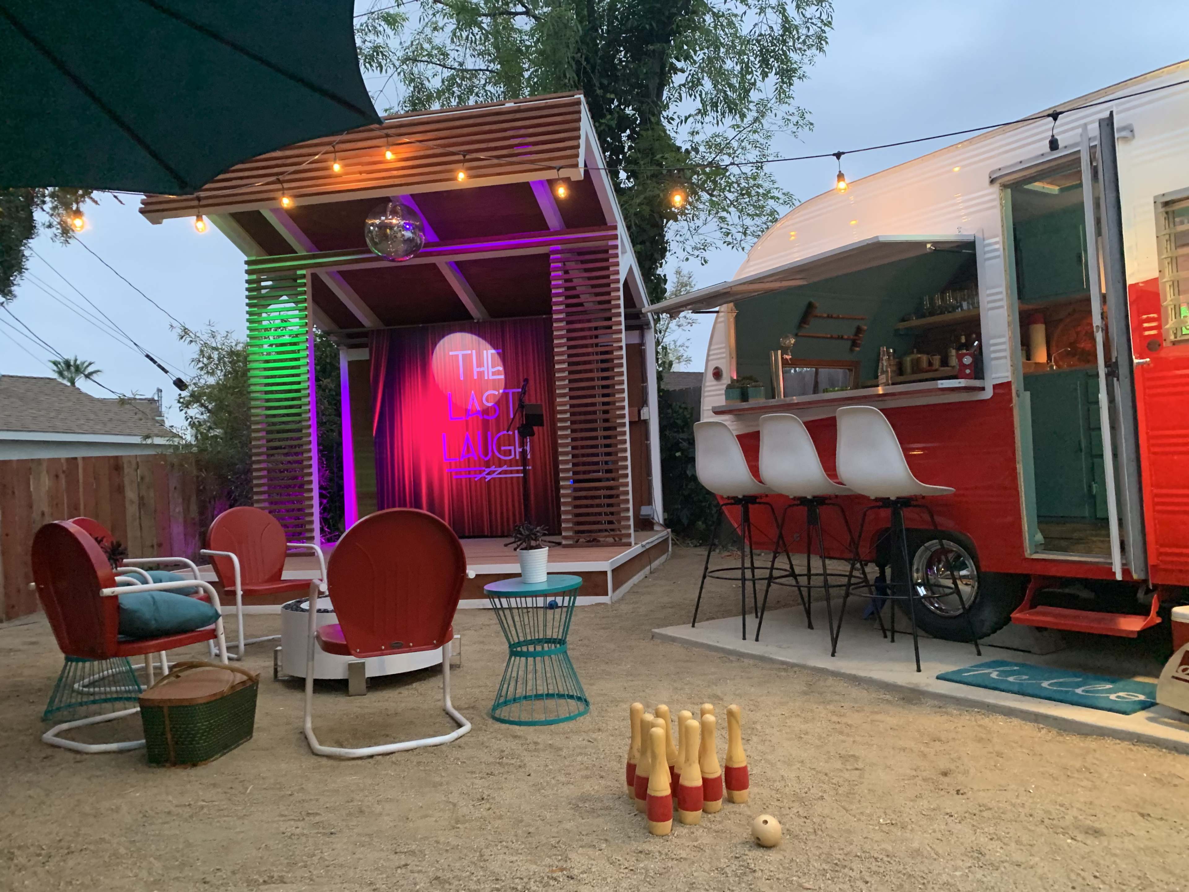 A cozy outdoor space featuring a retro trailer, a small stage with a colorful sign, and a bowling setup on the ground.