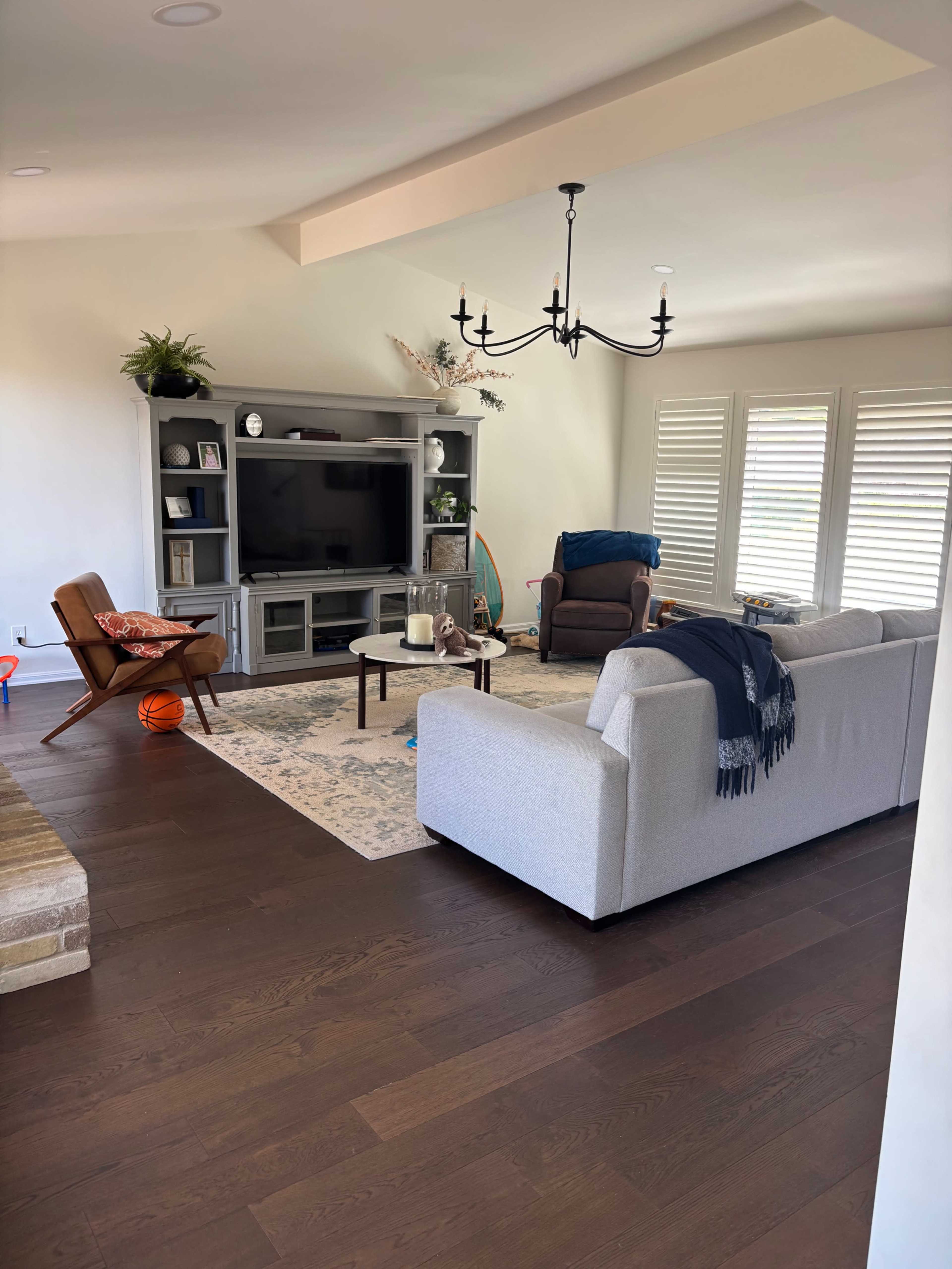 The image shows a living room with a cozy seating area, a television cabinet, and large windows featuring plantation shutters.