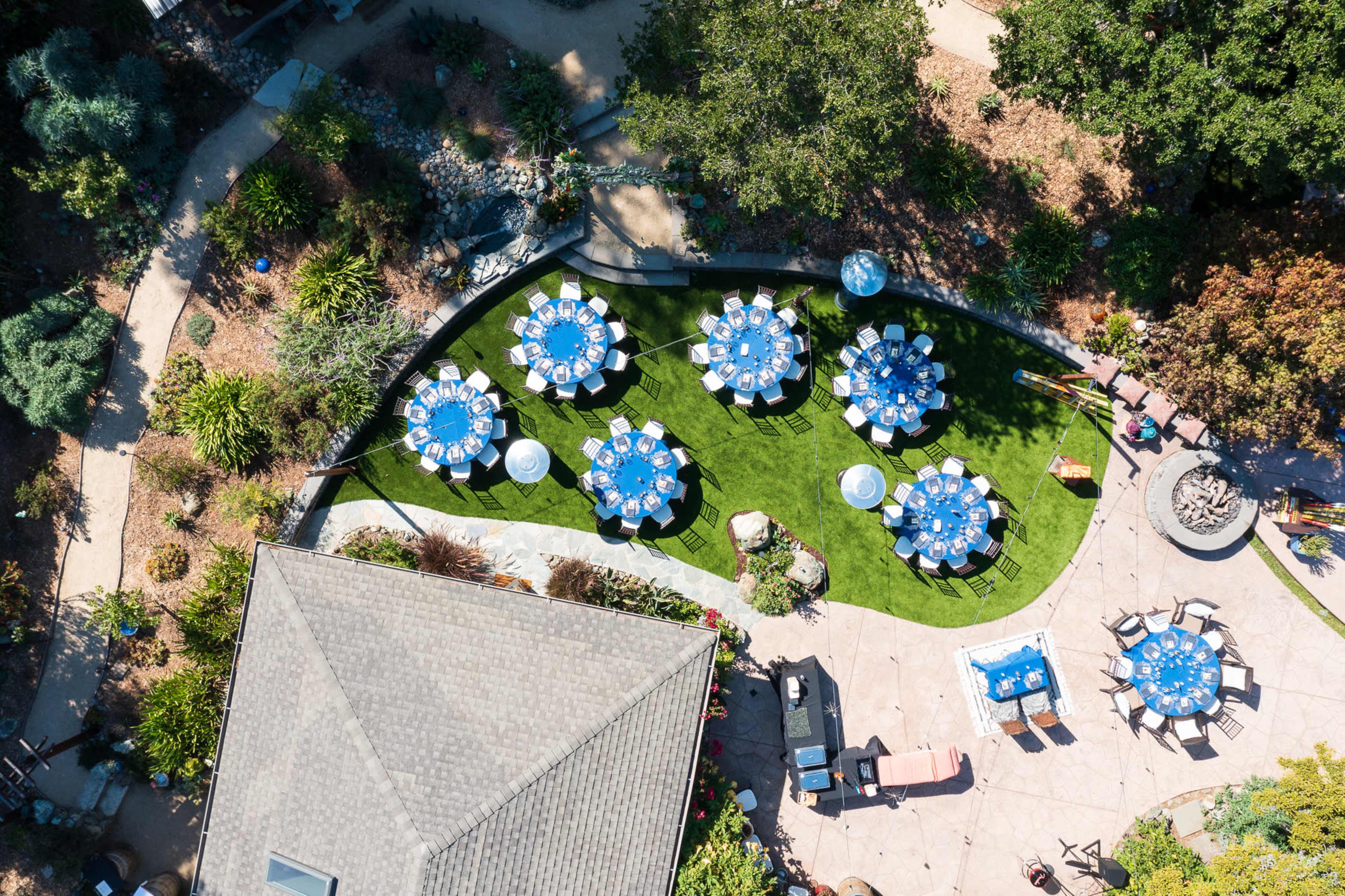 A landscaped outdoor area with several round tables set with blue tablecloths arranged on a green lawn.