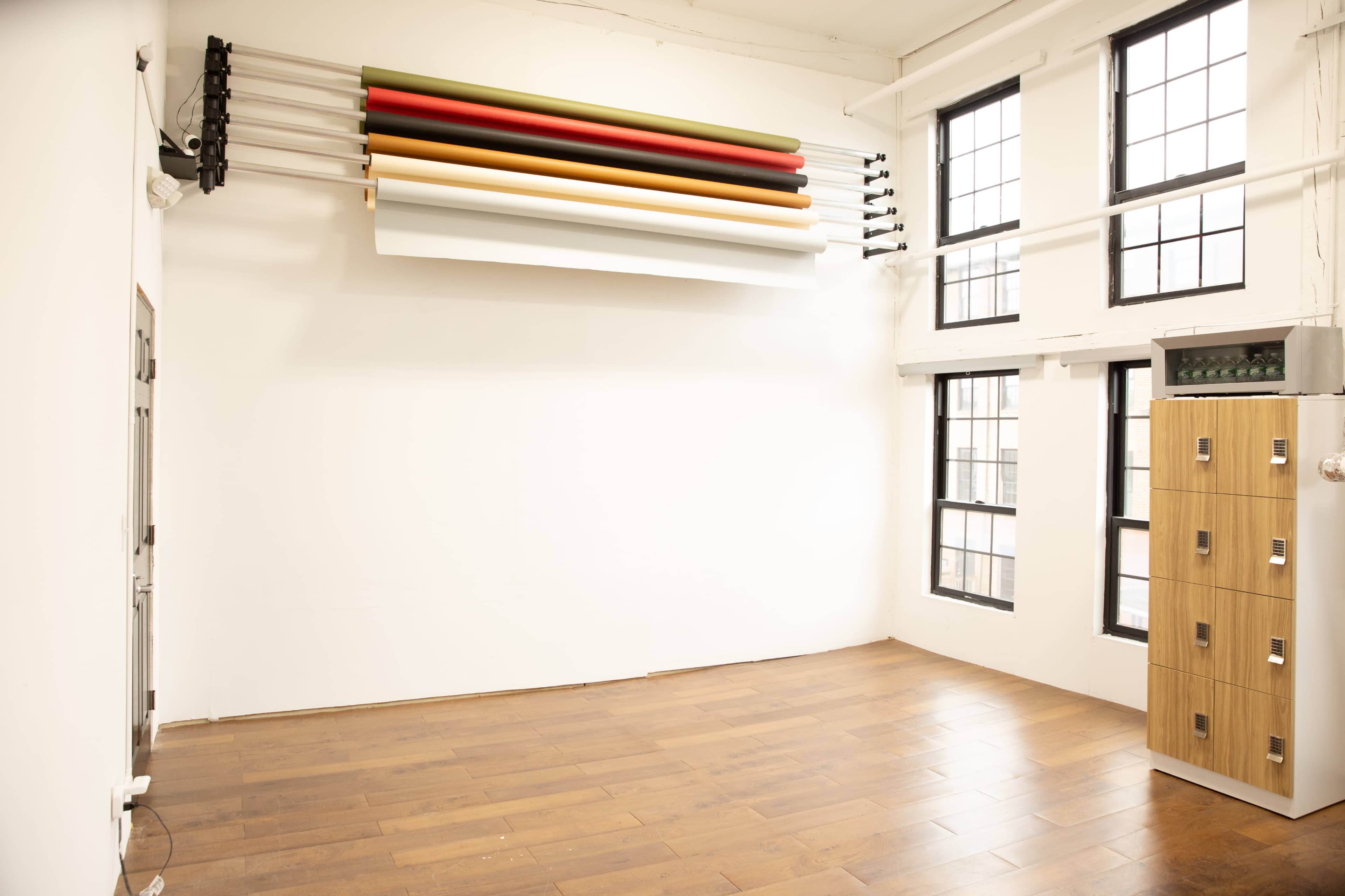 The image shows a spacious, empty room with wooden flooring, large windows, and a wall-mounted rack holding rolls of colored paper above a wooden cabinet.