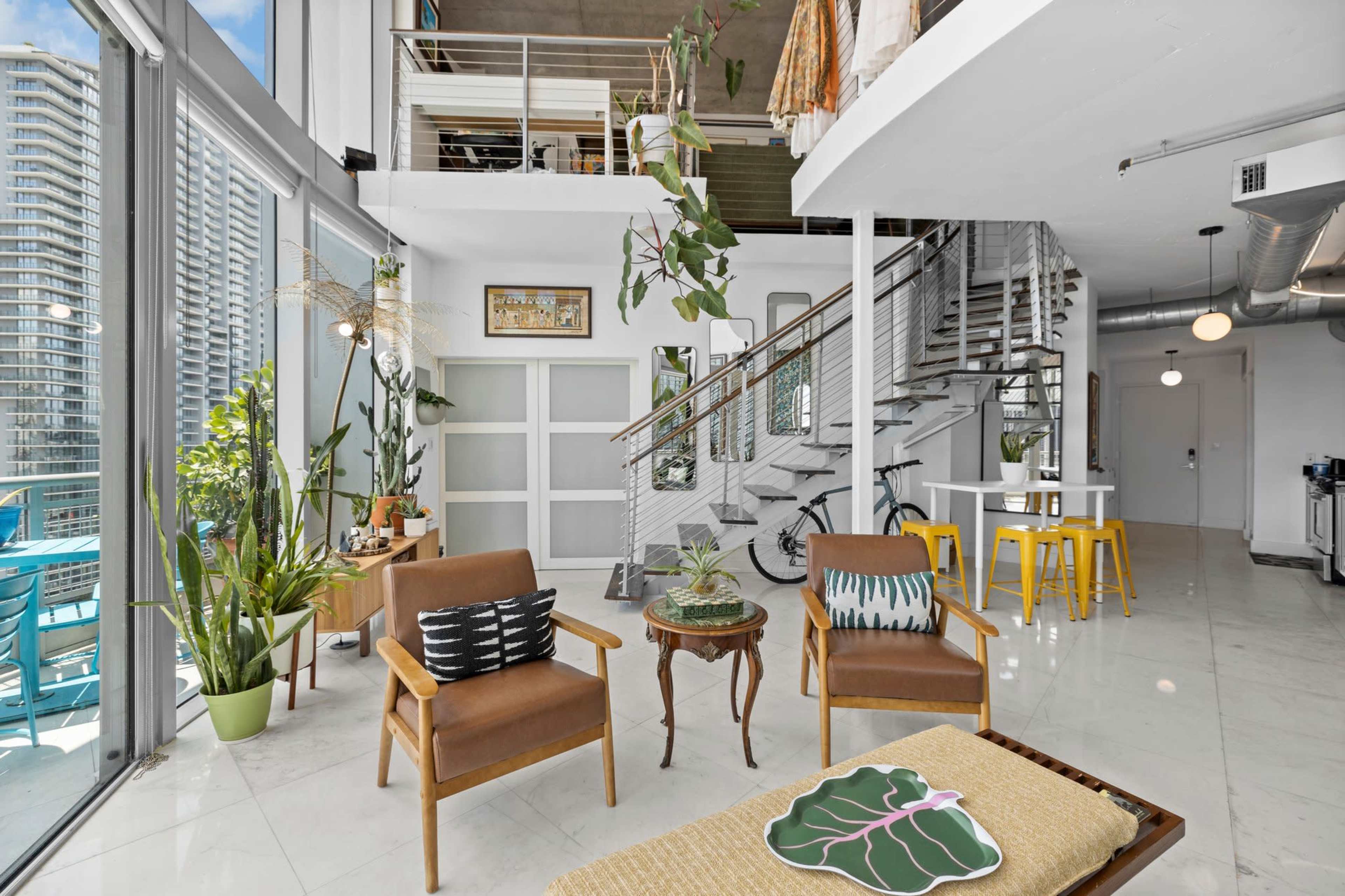 A spacious, modern living area with large windows, a staircase, and a mix of plants, furniture, and decor elements.