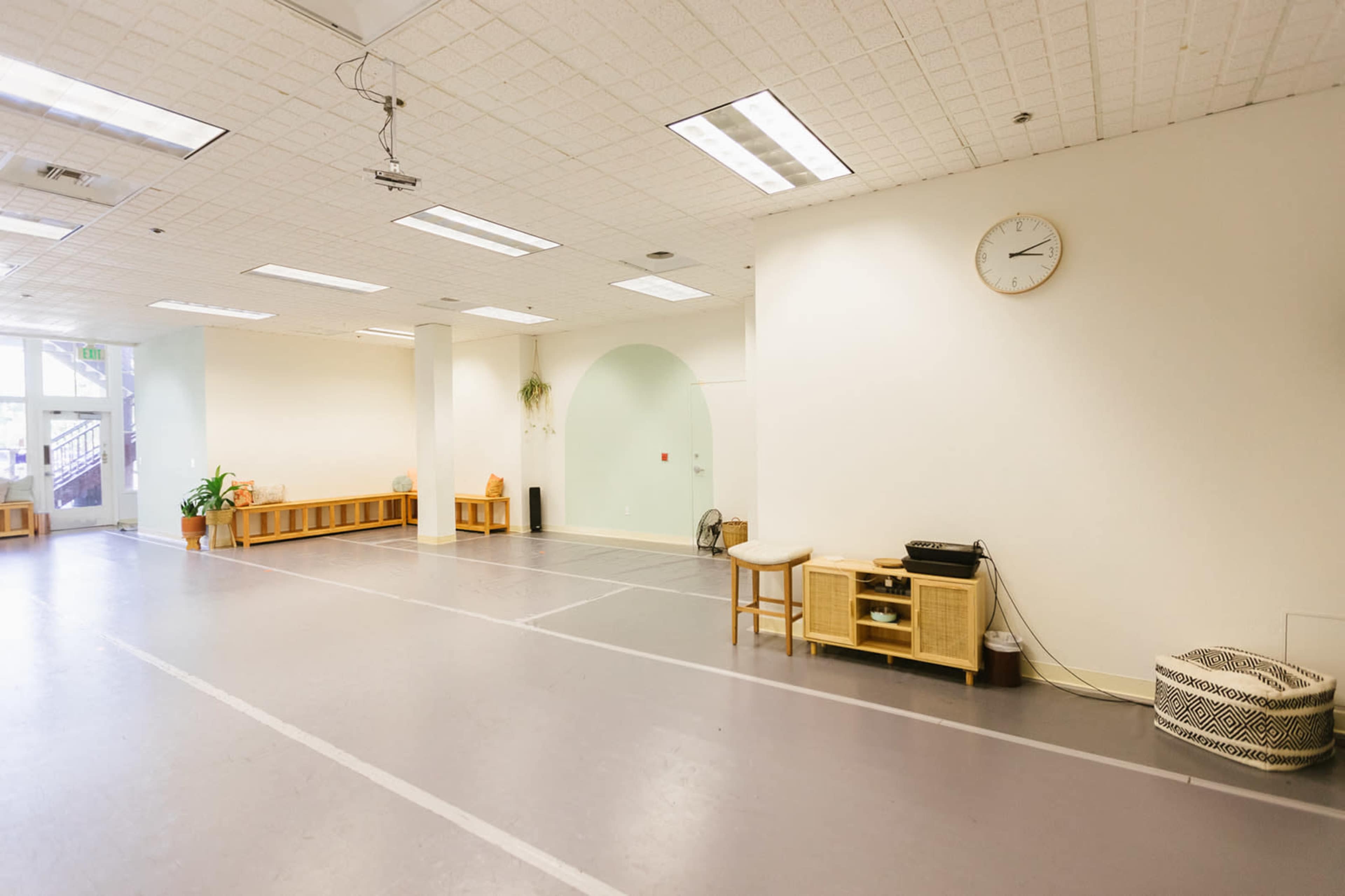 A spacious, empty room with light gray flooring, a clock on the wall, and minimal furniture along the sides.