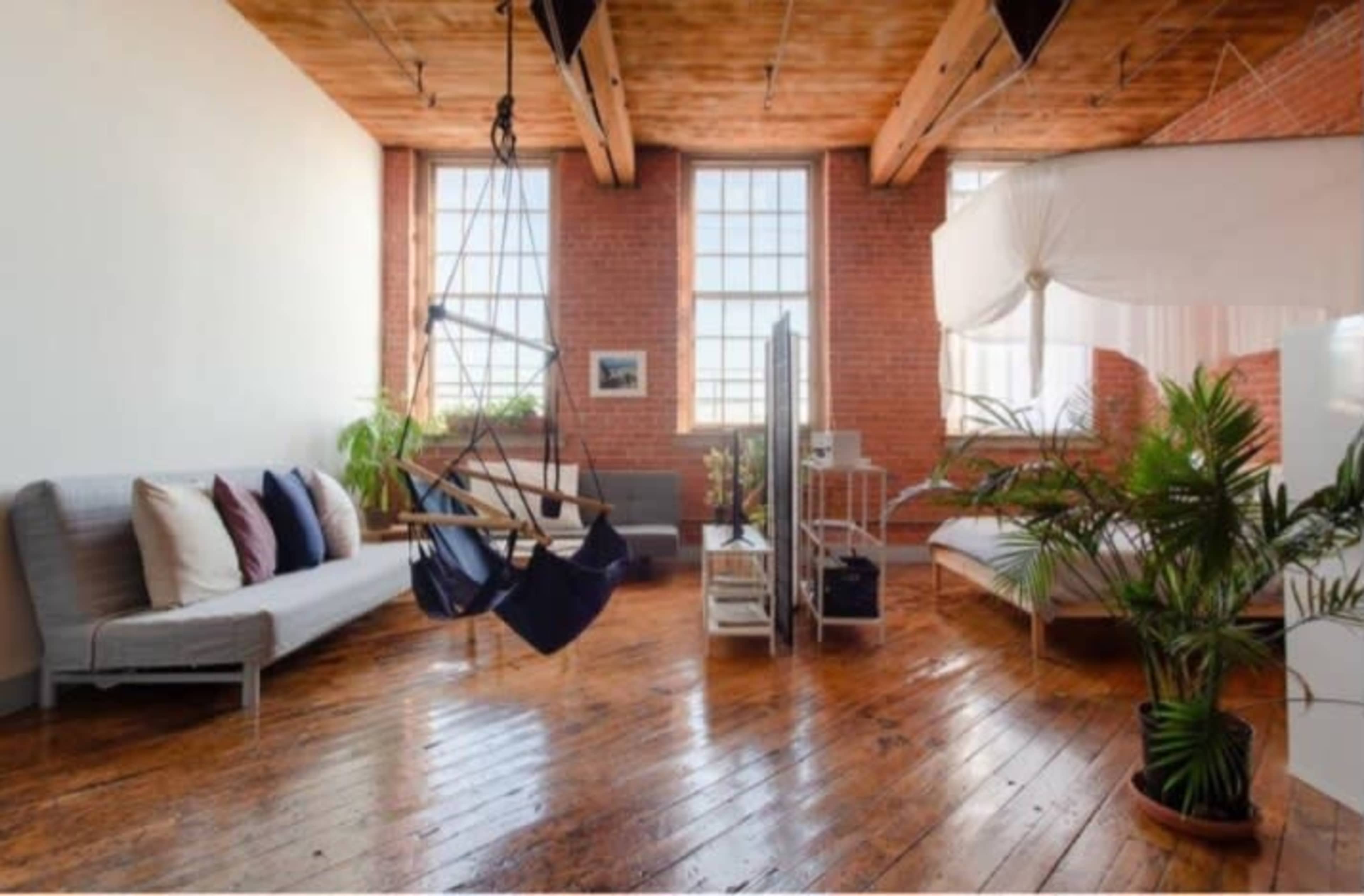 The image shows a spacious room with hardwood floors, large windows, a swing chair, and plants, featuring modern furniture against a brick wall.
