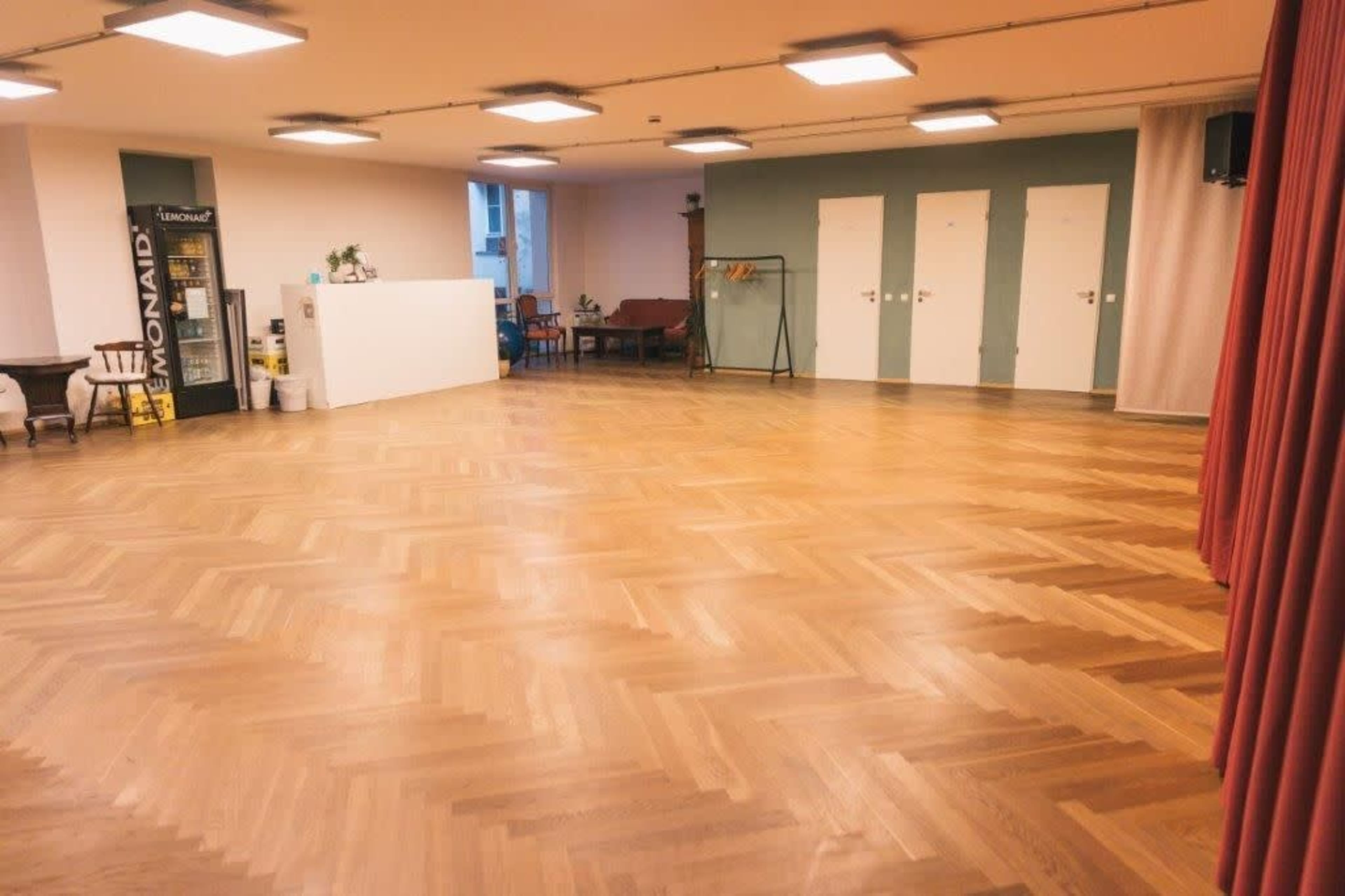 The image shows a spacious room with wooden flooring, featuring a white partition, a seating area with a table, and several doors on one wall.