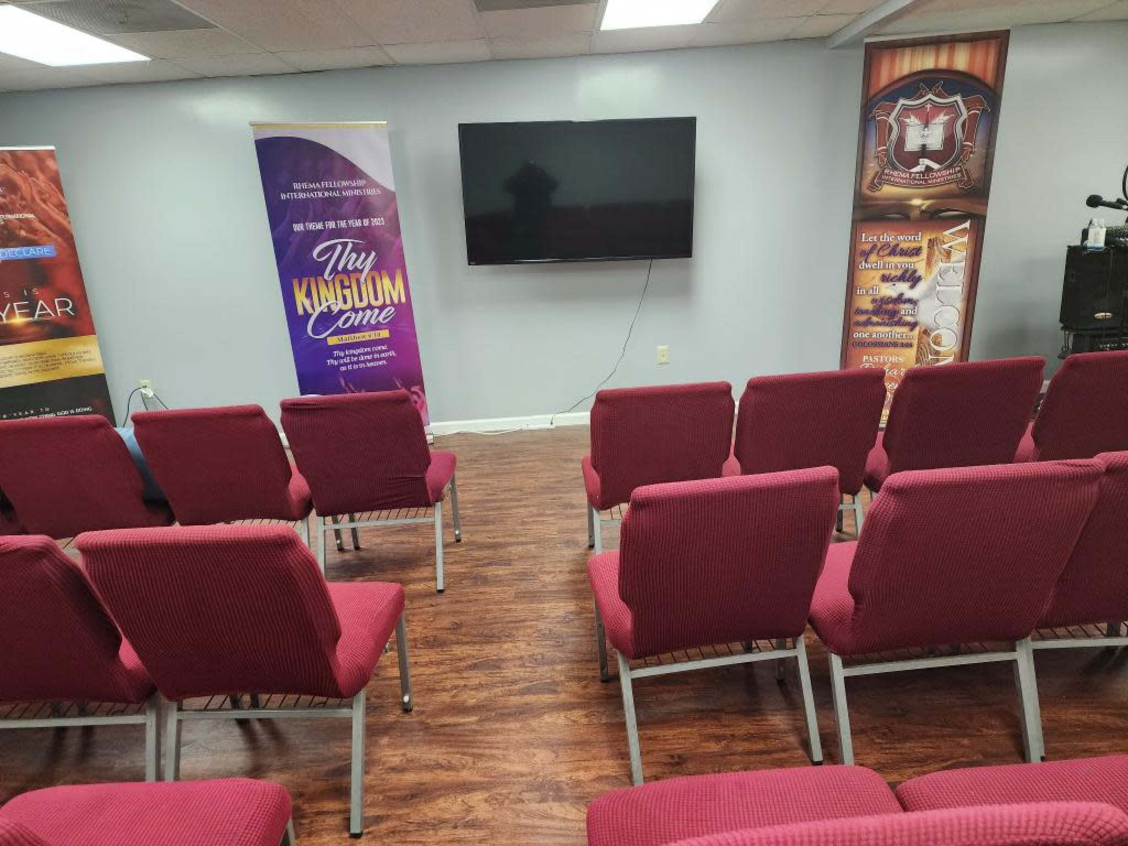 A small meeting room with rows of red chairs facing a wall-mounted television and several promotional banners.