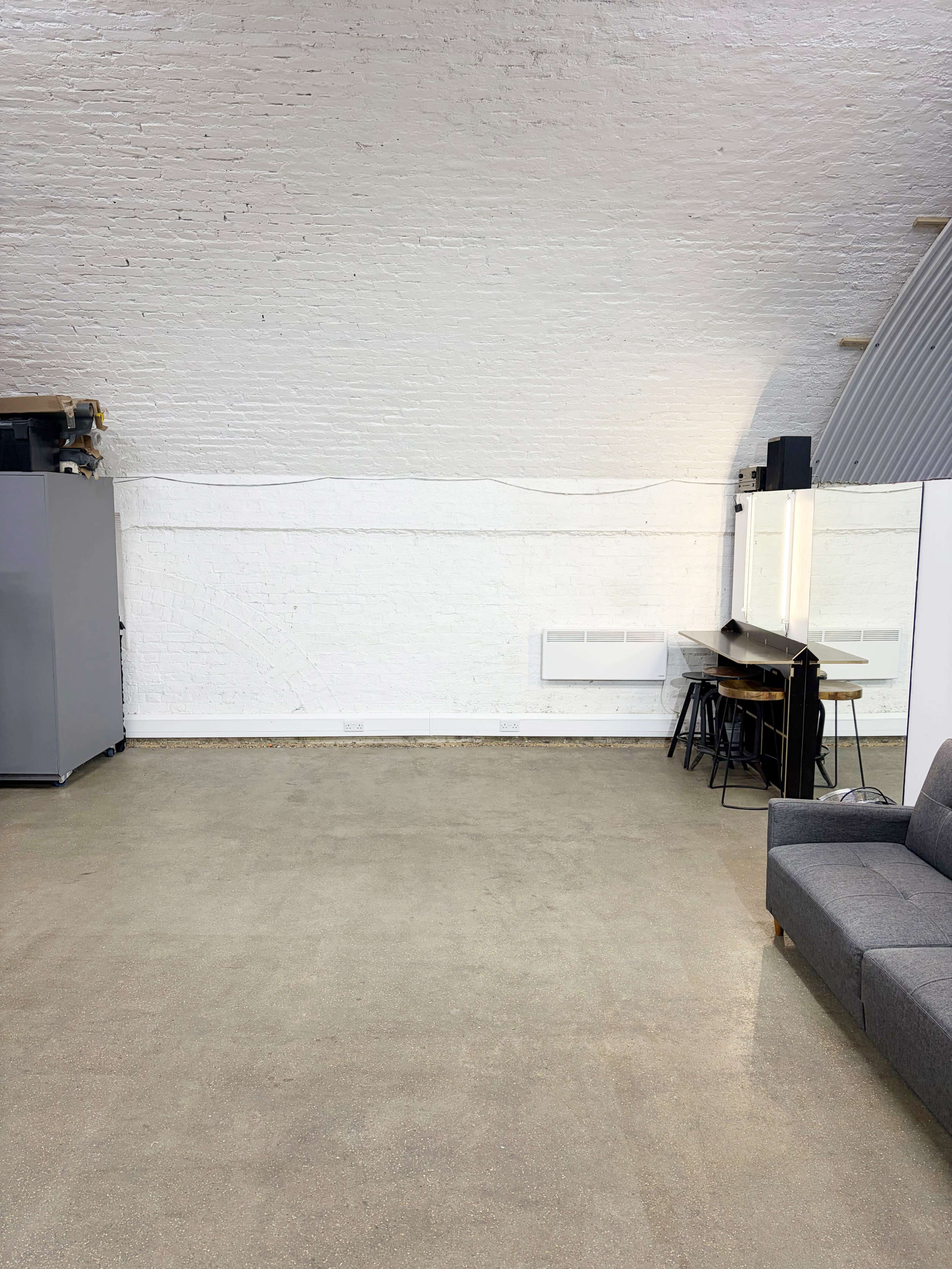 The image shows an empty space with a smooth concrete floor, a gray sofa, a mirrored wall, and a row of black chairs beside a table against a white-brick backdrop.
