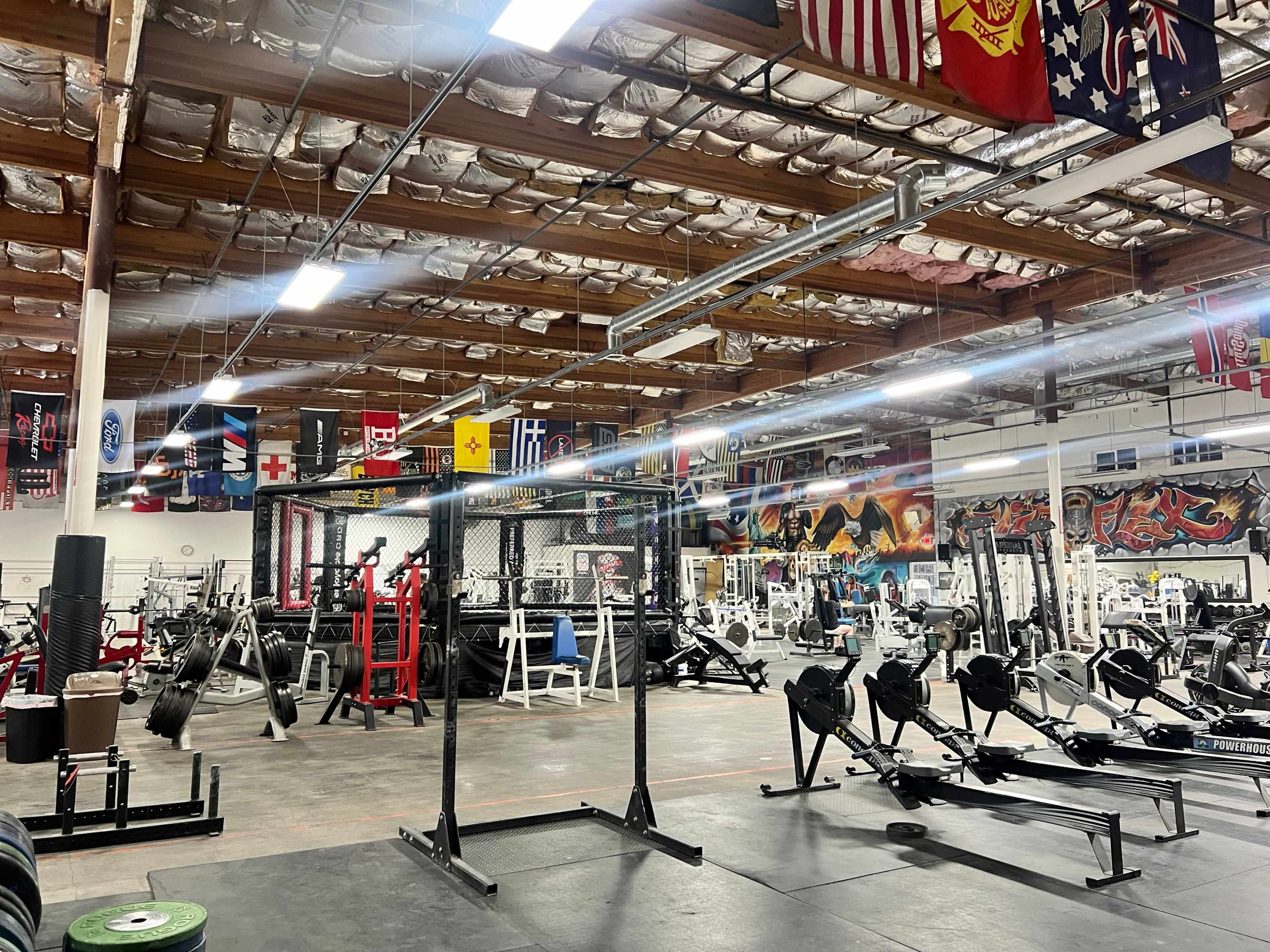 A spacious gym filled with various weightlifting equipment and workout machines, decorated with flags and murals on the walls.