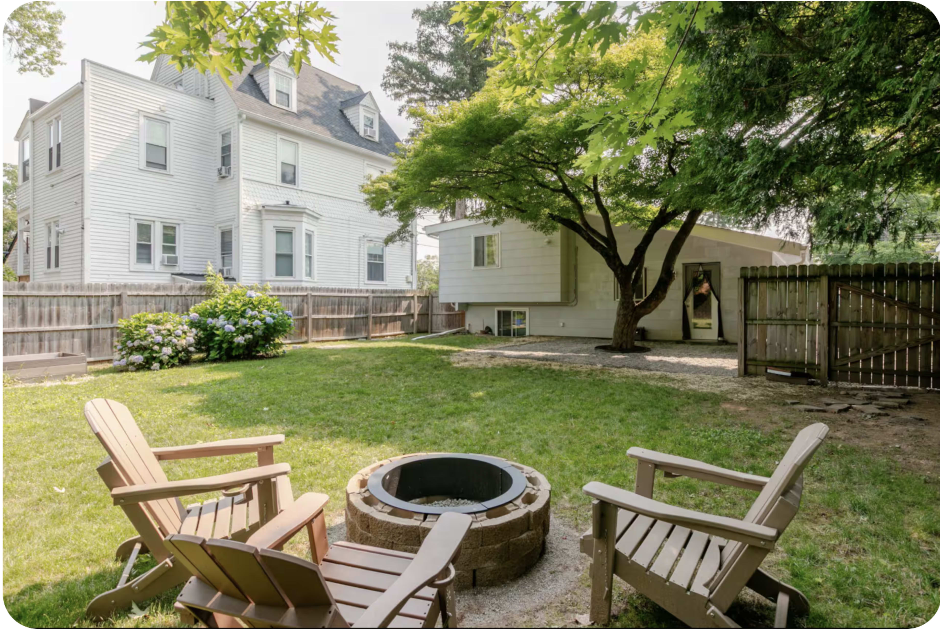 The image shows a backyard with a fire pit surrounded by wooden chairs, green grass, and trees, with two houses visible in the background.