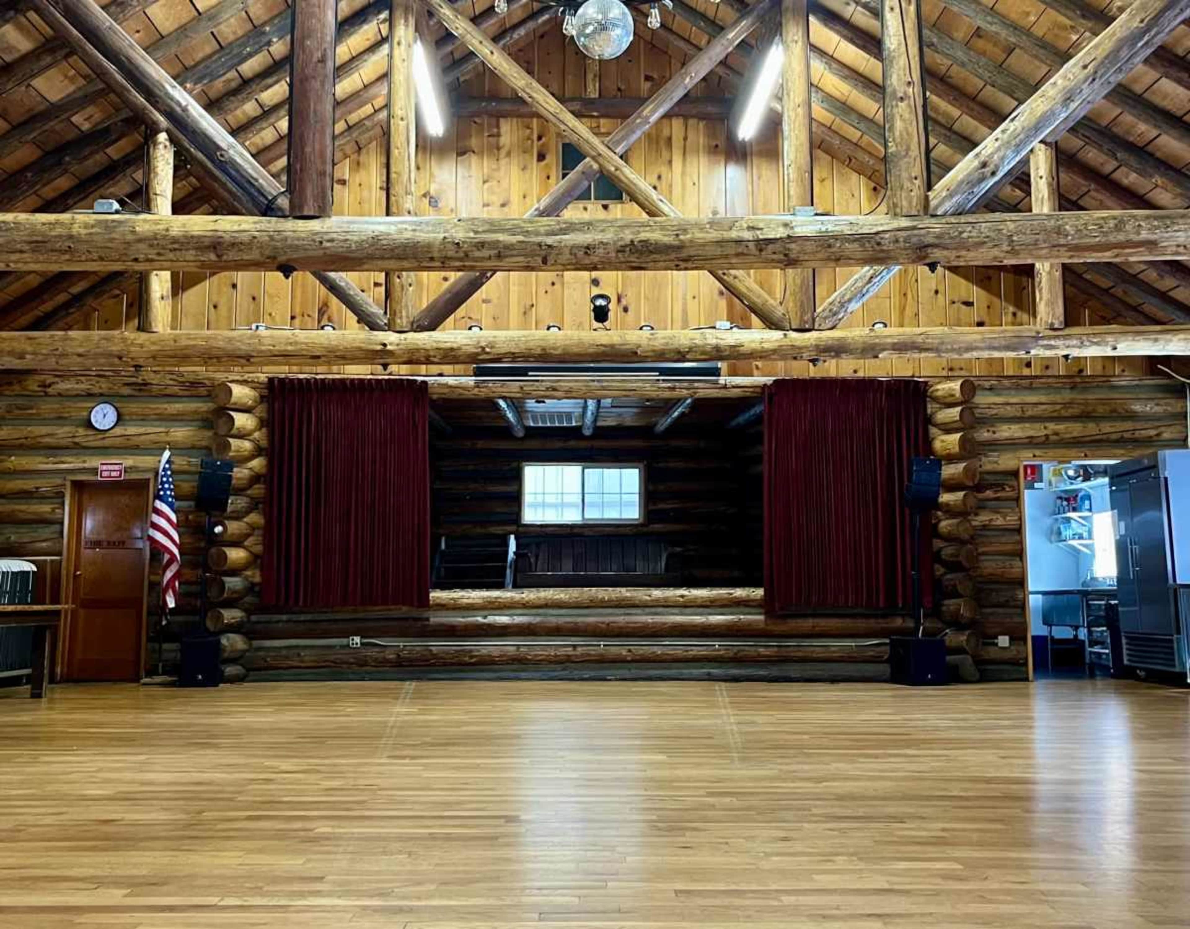 The image shows a spacious interior of a rustic hall with wooden log walls, a wooden floor, and a stage flanked by red curtains.