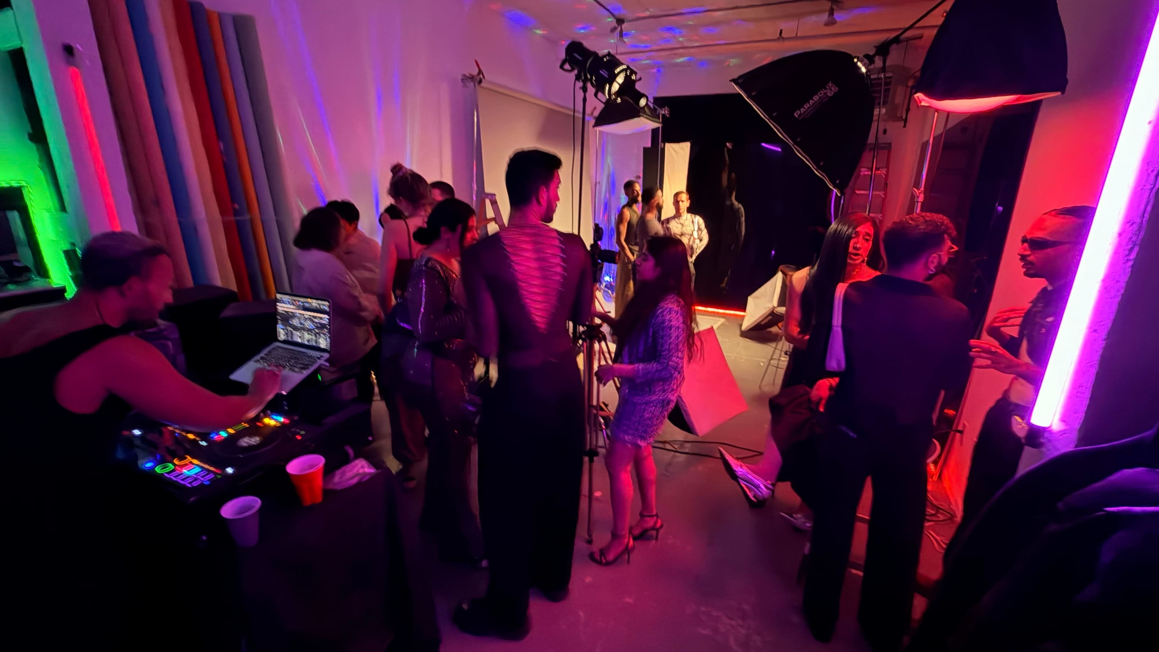 A crowded studio space features individuals socializing and working behind the scenes of a photoshoot, with colorful lights illuminating the area.
