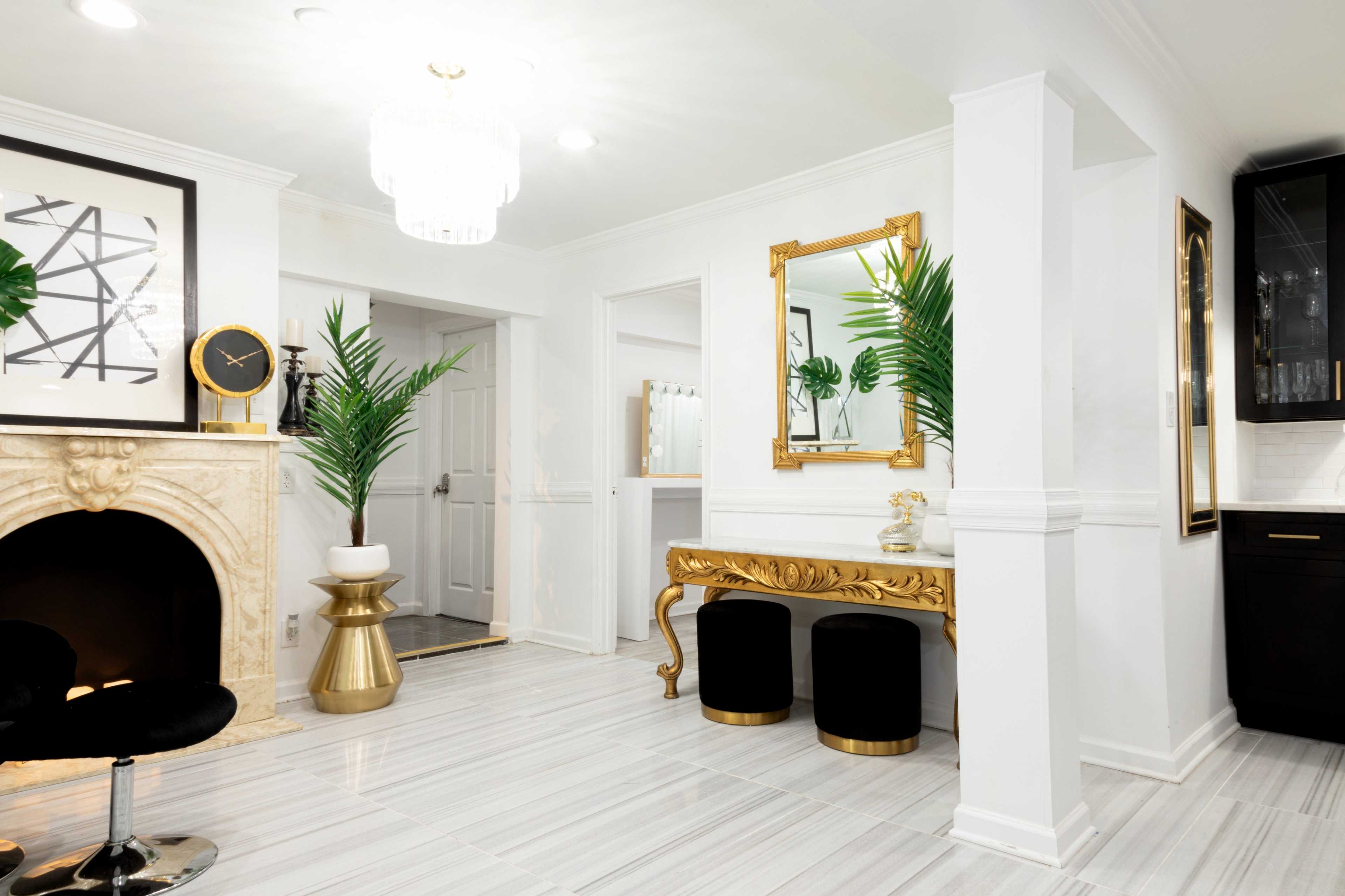 A brightly lit foyer with a gold-framed mirror, a decorative table, black stools, a faux plant, and a fireplace with a marble surround.