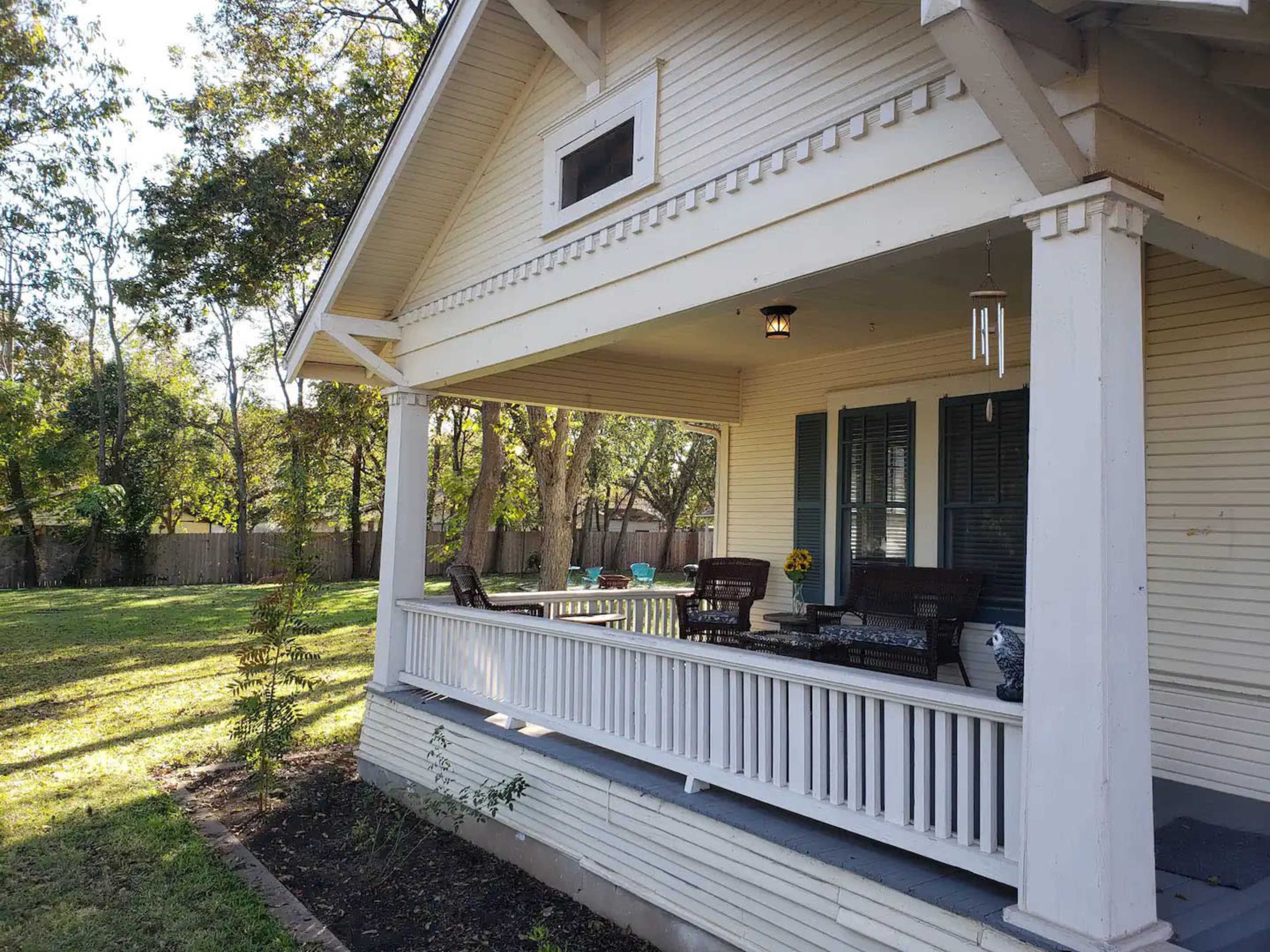 A single-story house with a front porch, featuring wicker furniture and a flower vase, surrounded by a grassy yard and trees in the background.