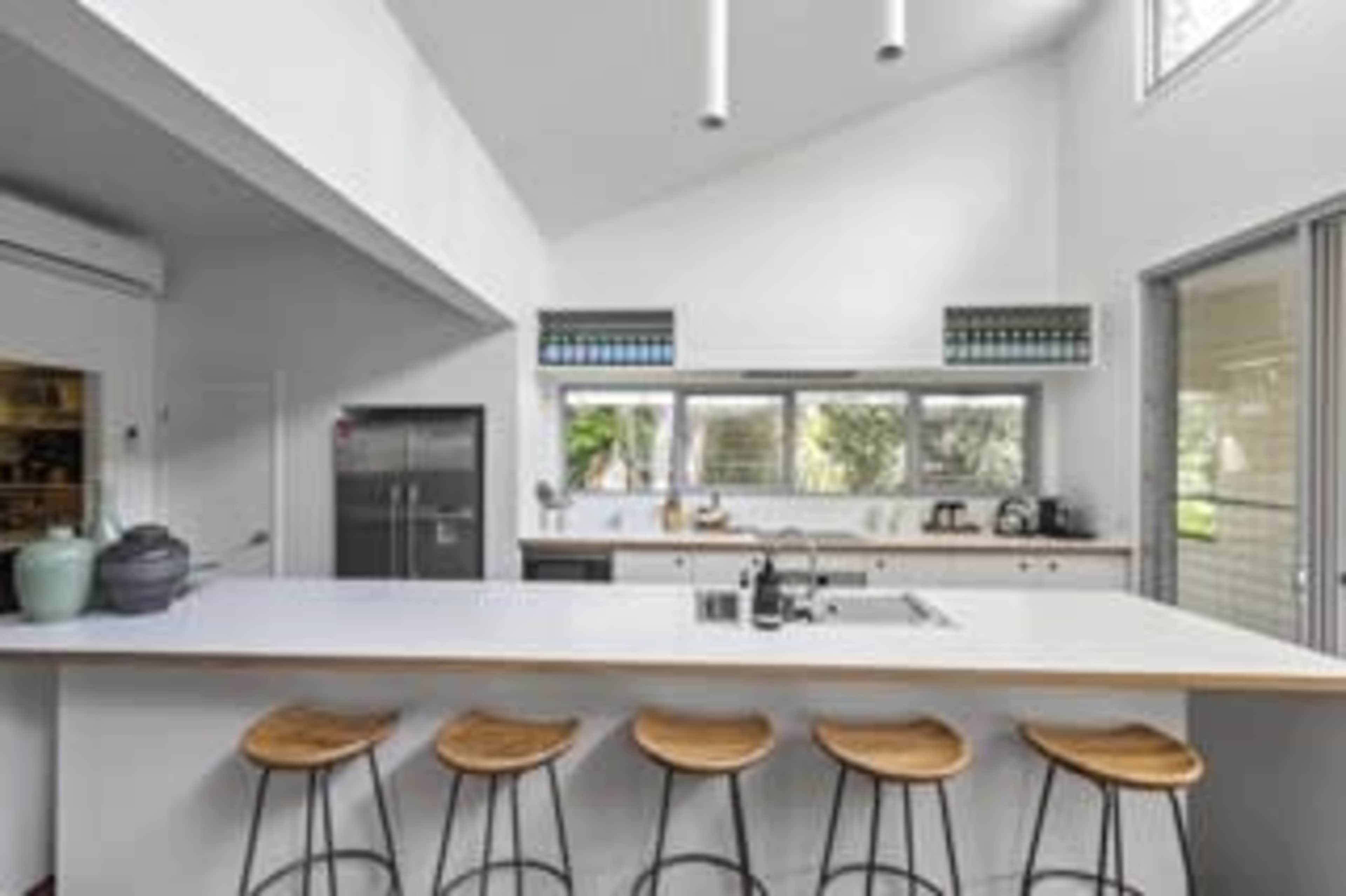 A modern kitchen features a large island with four wooden stools, sleek appliances, and large windows allowing natural light.