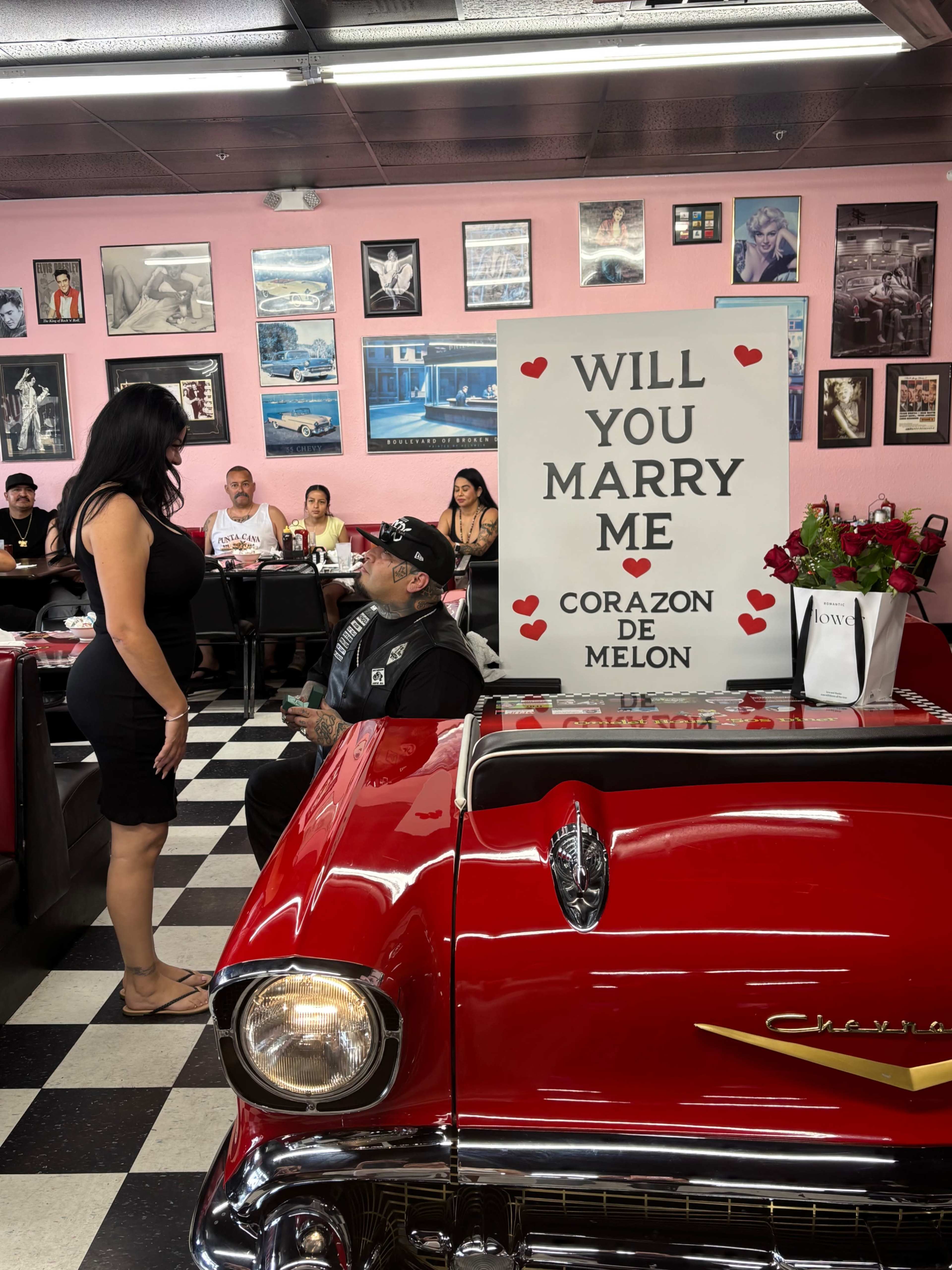 A man sitting in a vintage red car in a diner is proposing to a woman standing in front of him, with a sign that says "WILL YOU MARRY ME" decorated with hearts.