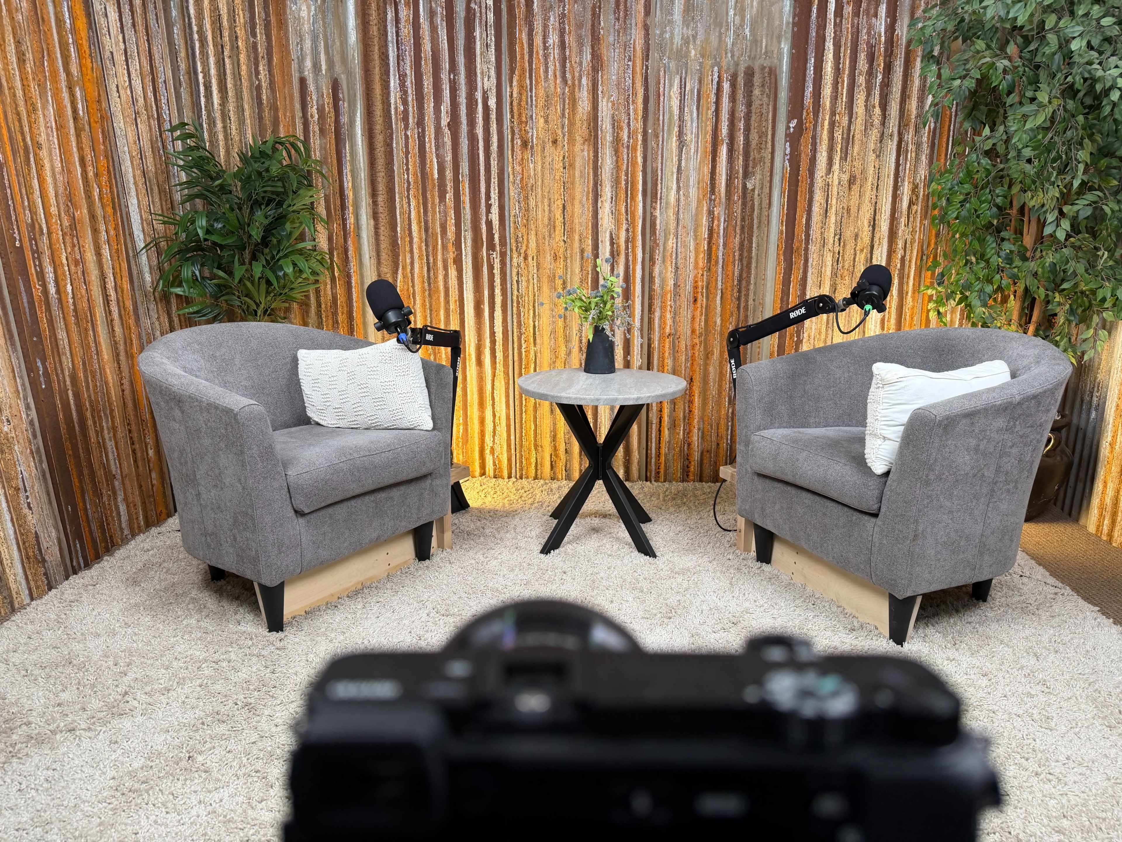 The image shows a cozy podcast studio setup with two gray armchairs facing a small black table, surrounded by a rustic wooden backdrop and potted plants.