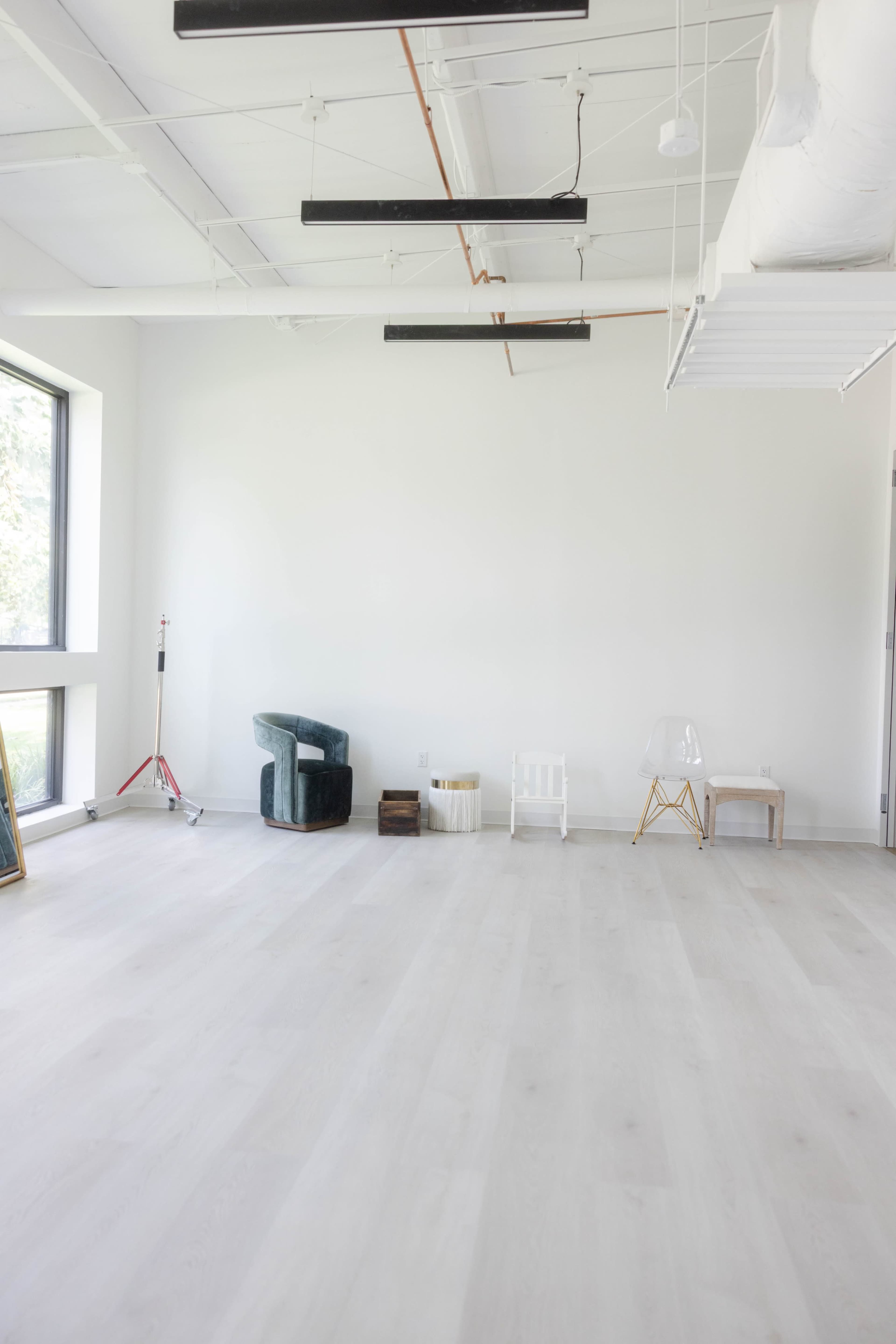 A bright, empty room features light-colored wooden flooring, large windows, and minimal furniture, including a green armchair and a clear chair.