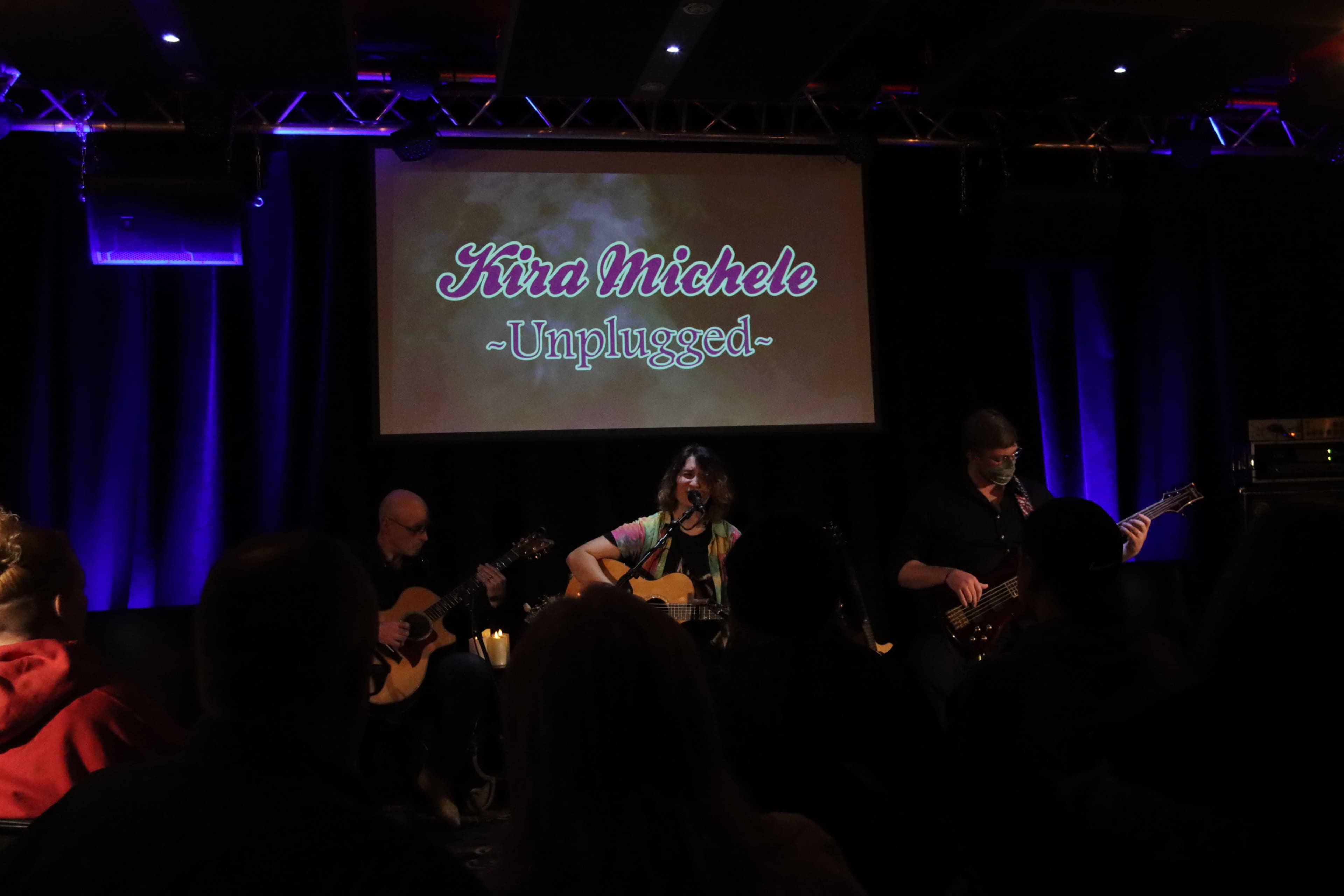 A woman performs on stage with two musicians accompanying her, while an audience listens in a dimly lit venue.
