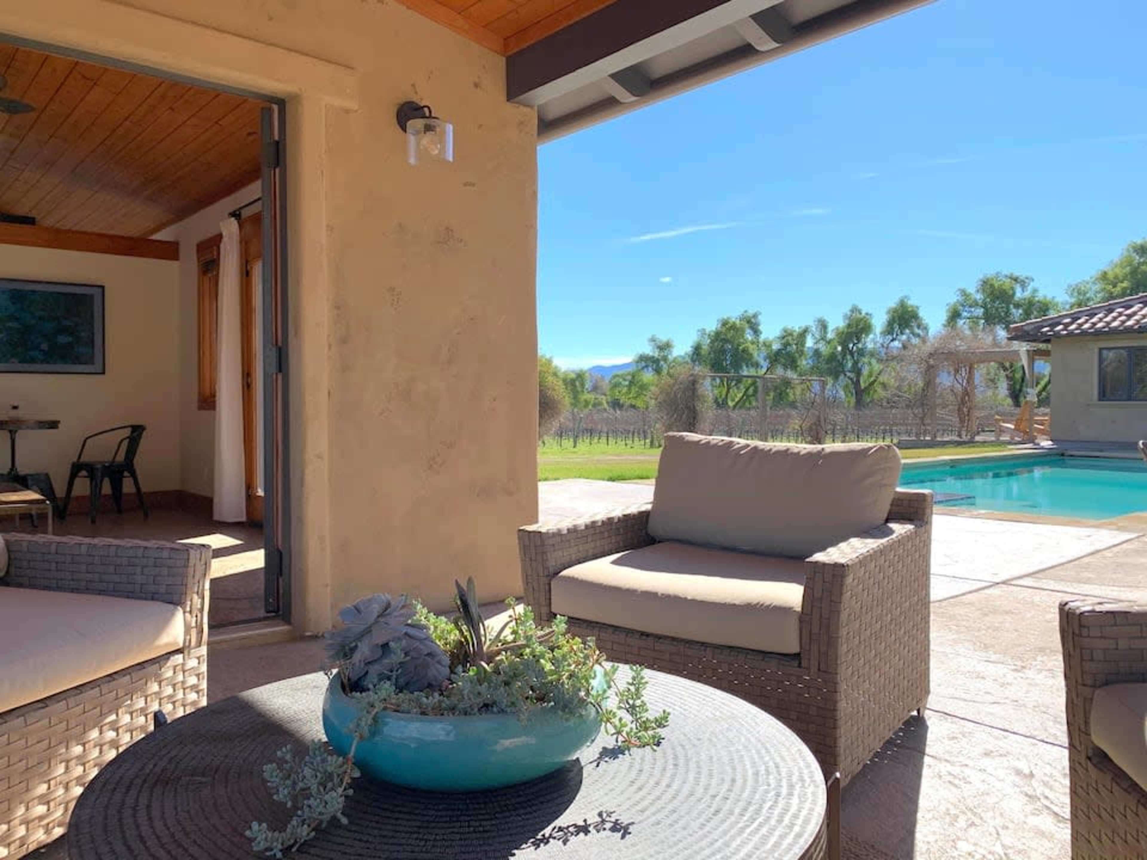 A patio area with wicker chairs and a table featuring a plant, overlooking a pool and a landscaped garden with trees in the background.