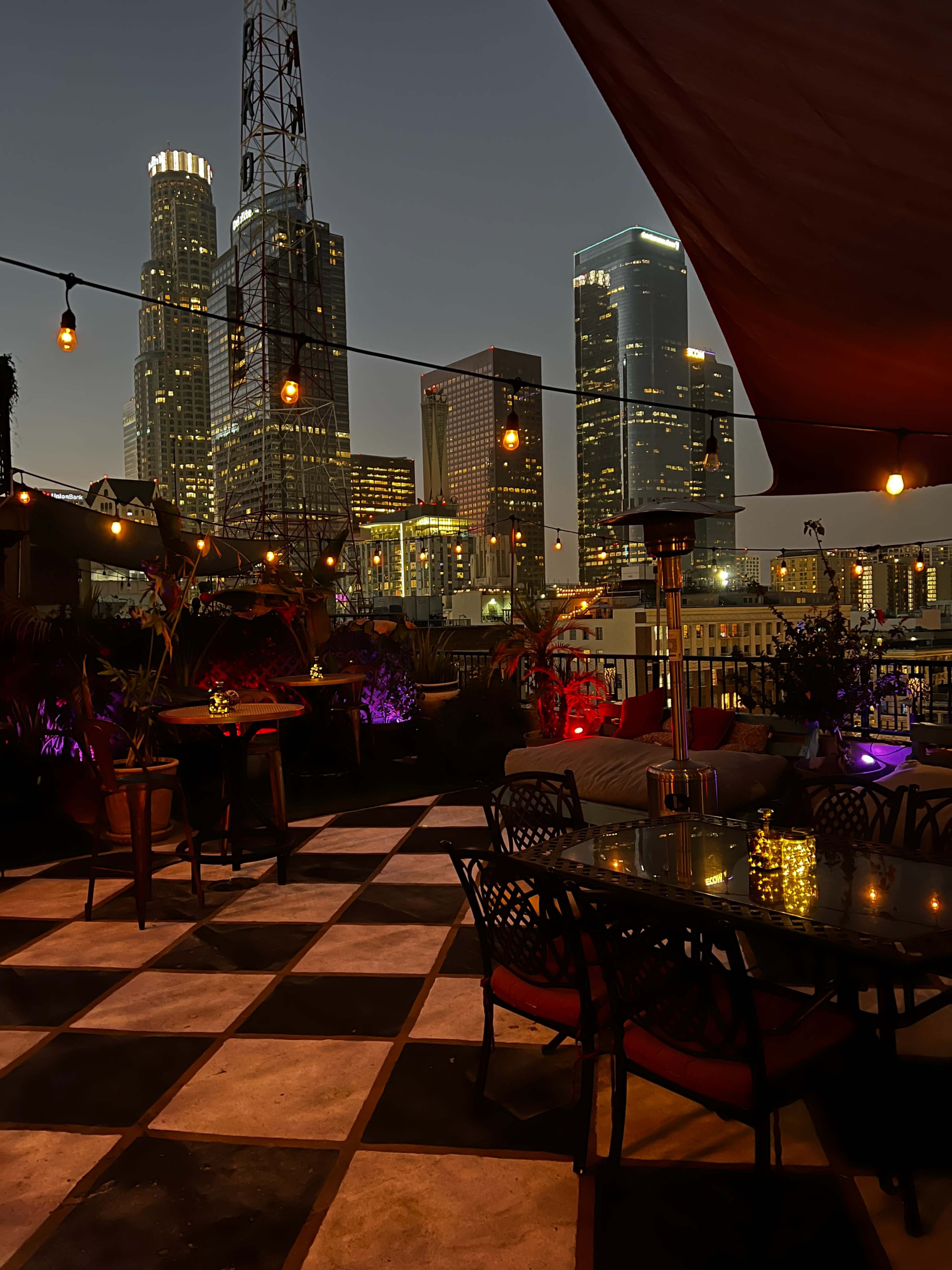 A rooftop terrace in Los Angeles at dusk, featuring a checkered floor, tables with illuminated centerpieces, and a backdrop of city skyscrapers.