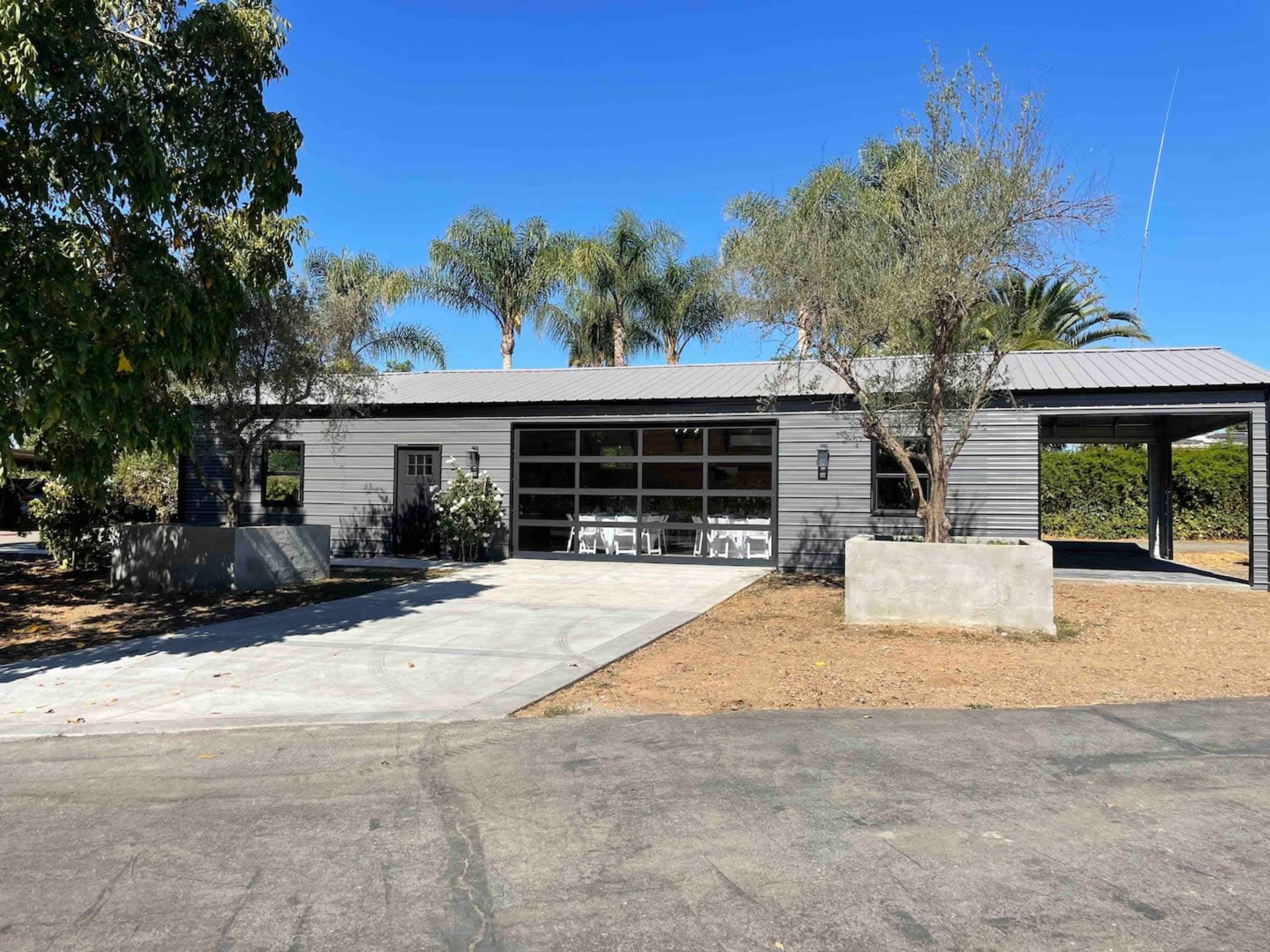 A modern single-story building with a garage door and a concrete driveway is surrounded by palm trees and landscaping.