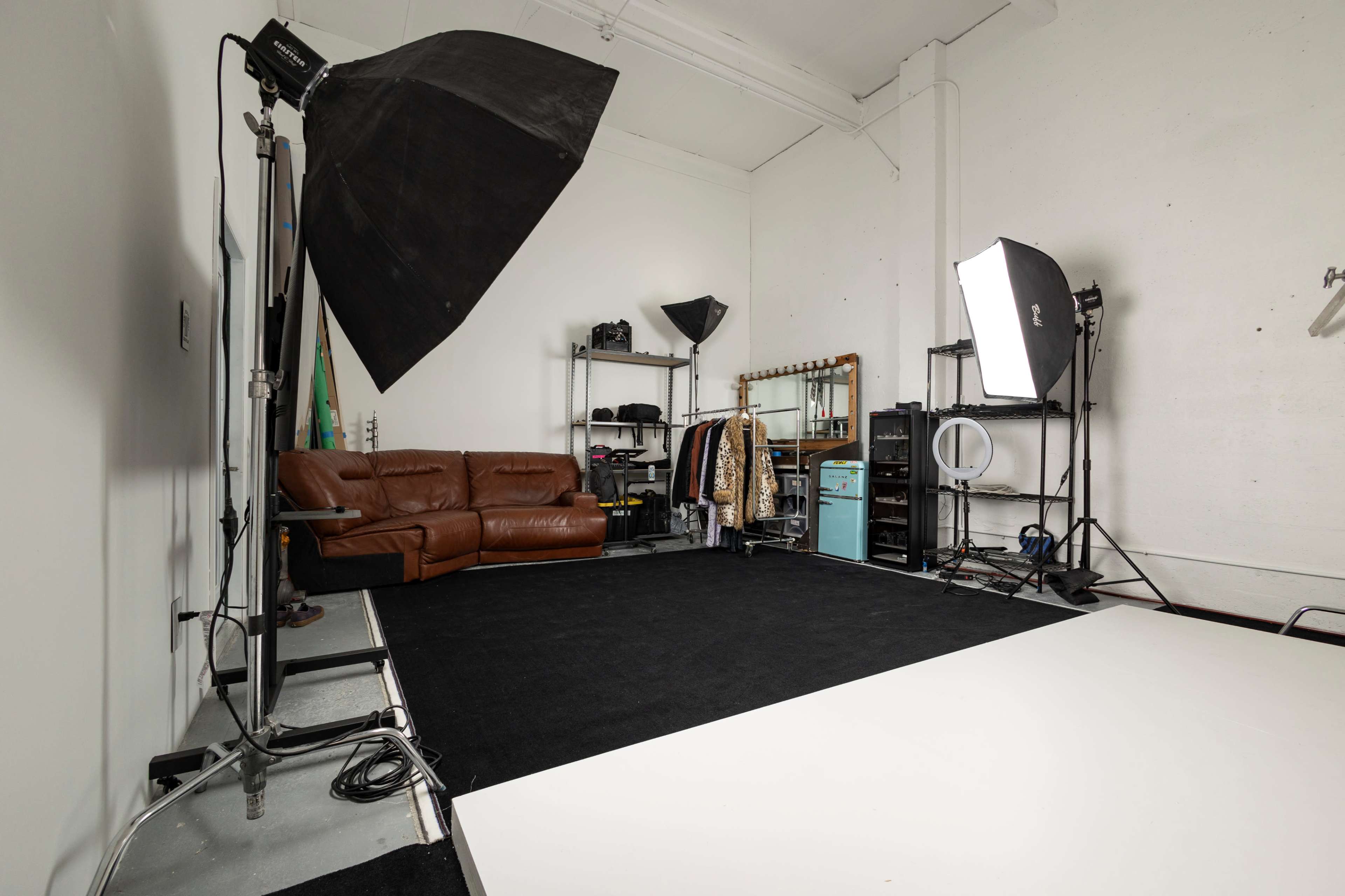 A photography studio with a brown leather couch, clothing racks, and various lighting equipment set up against white walls and a black carpet.