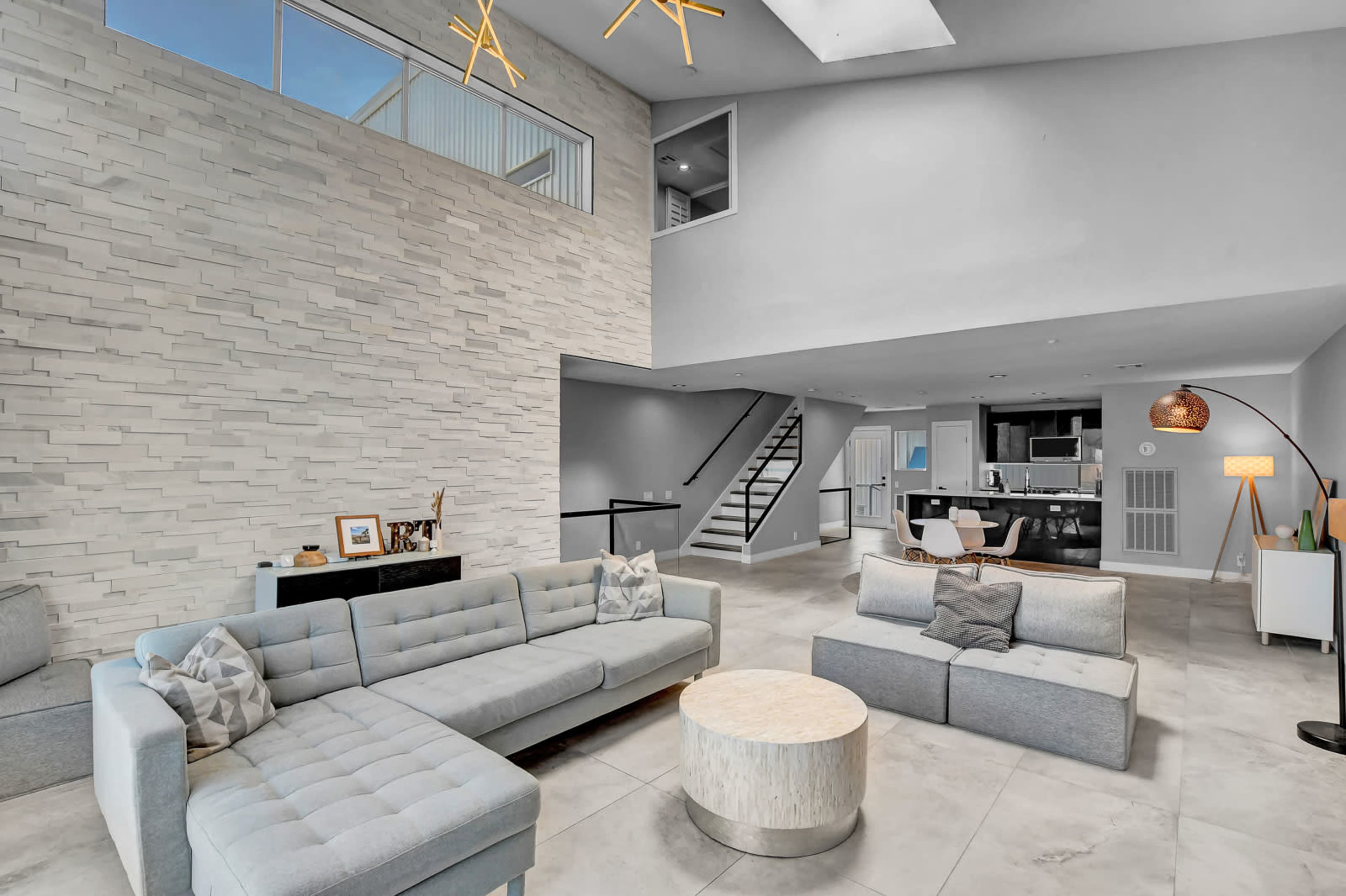 A modern living room features a high ceiling, contemporary furniture, and an open kitchen visible in the background.