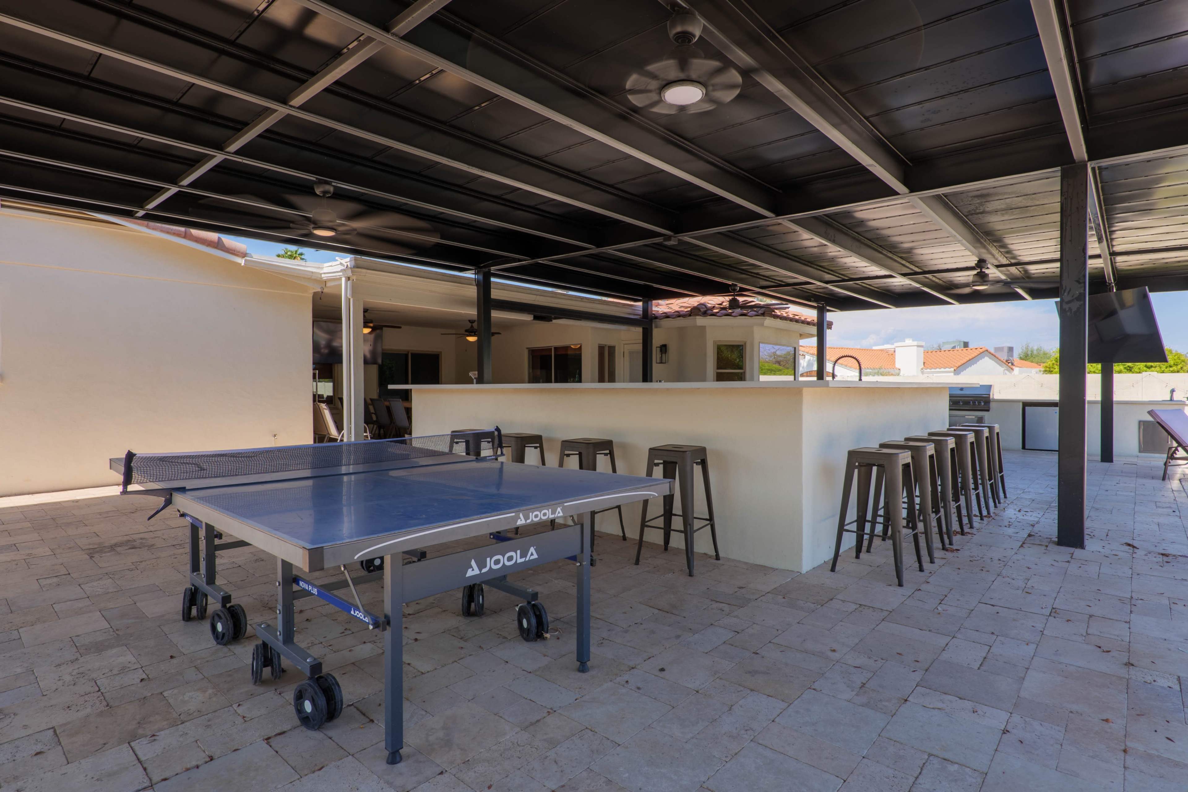 The image shows a covered outdoor area featuring a ping pong table and a bar with several stools.
