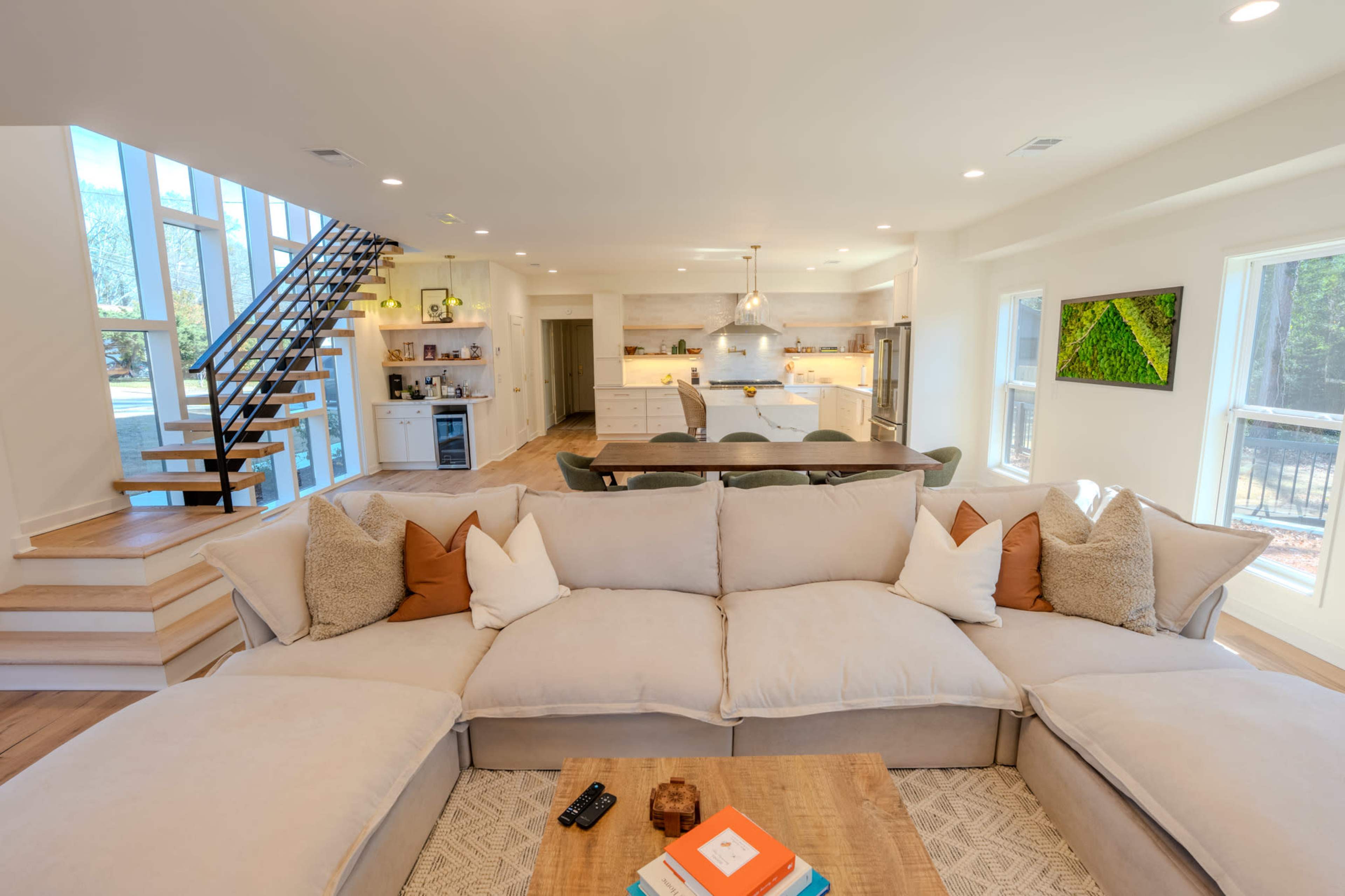 A spacious living room features a large sectional sofa with decorative pillows, facing a coffee table, while a staircase to the left leads to an upper level and the kitchen area is visible in the background.